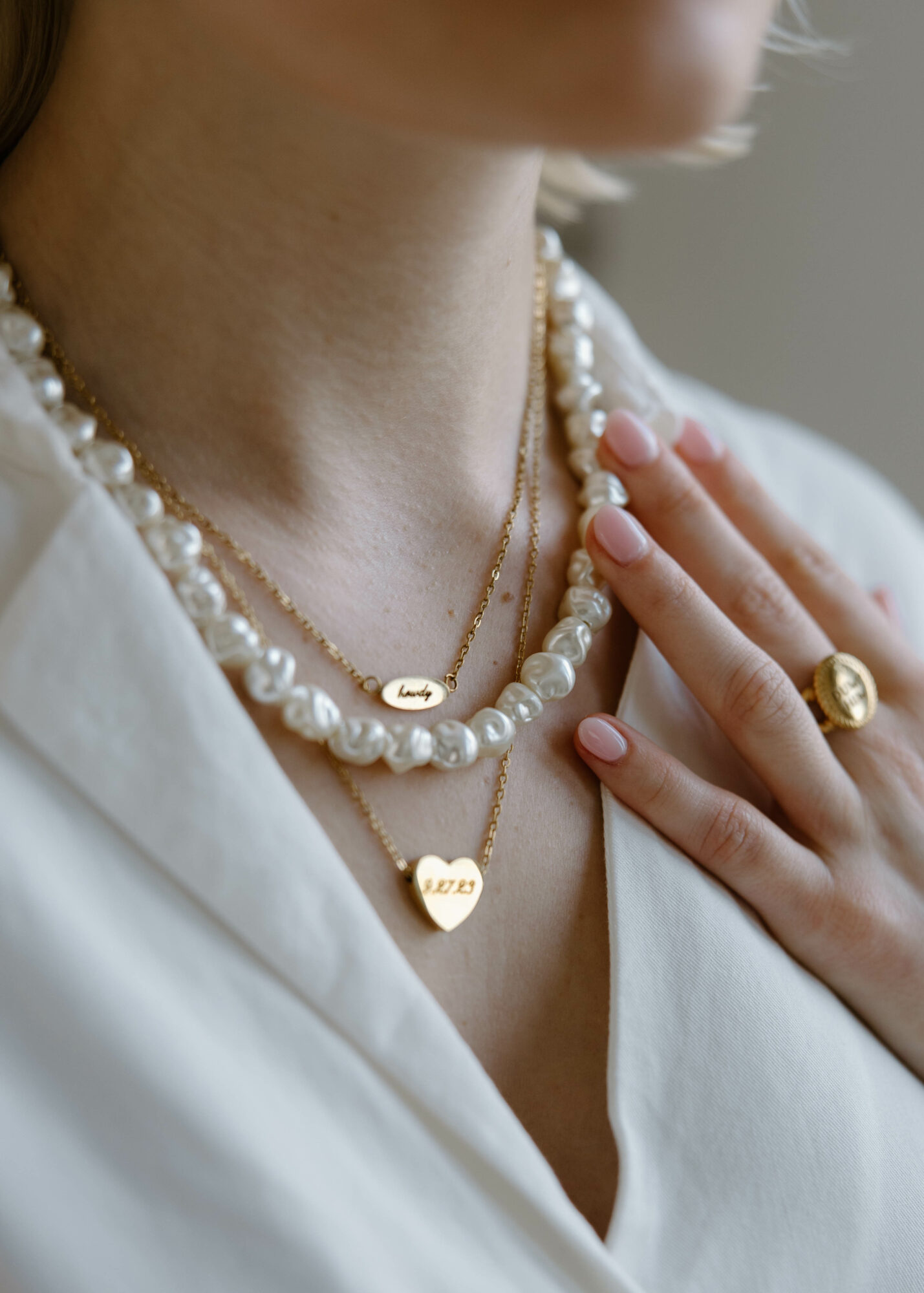 Close-up of a woman wearing layered pearl and gold necklaces, with a ring on her finger and hand on her chest.