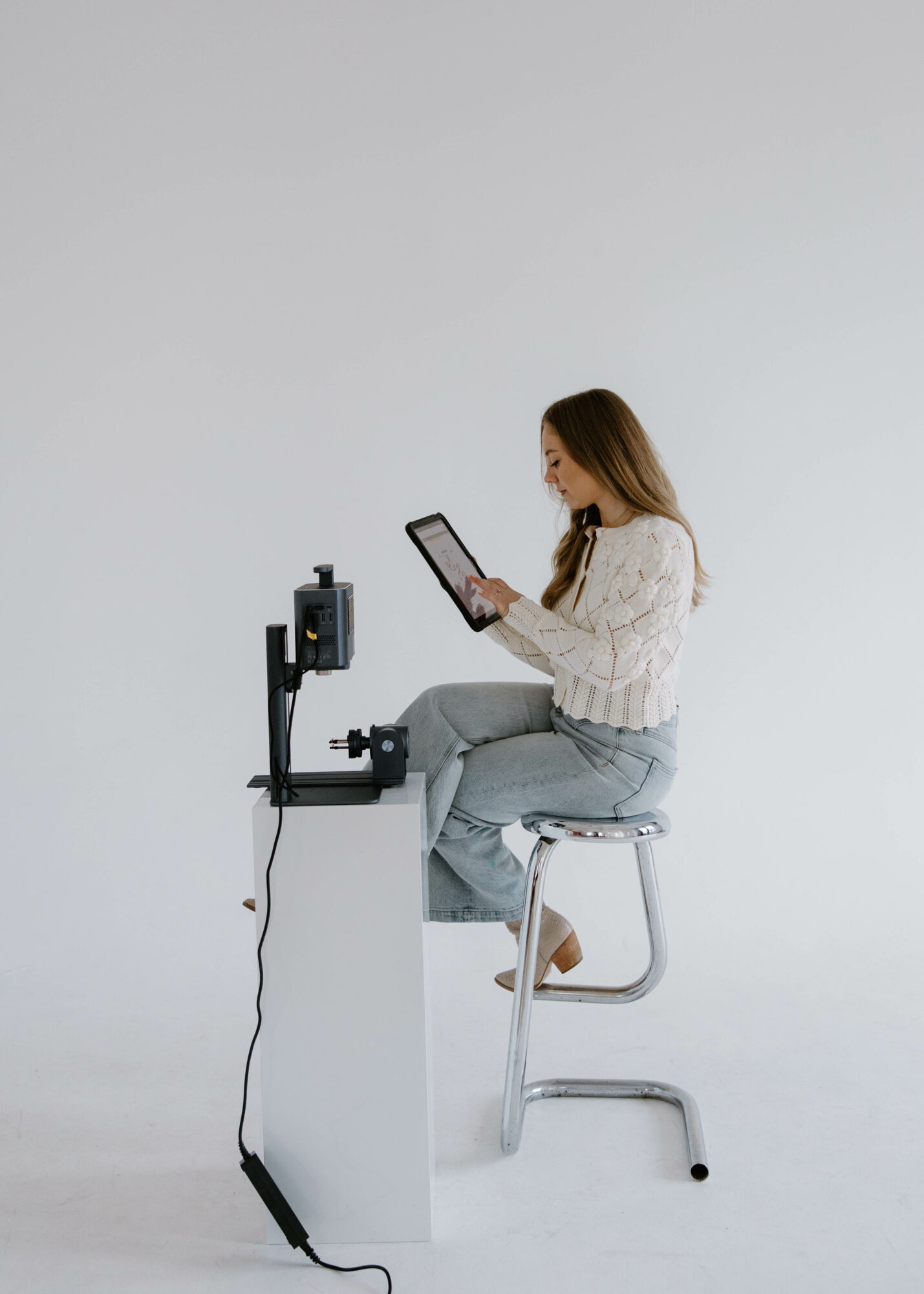 Woman sitting on a stool, using a tablet with a device on a white table, against a plain white background.