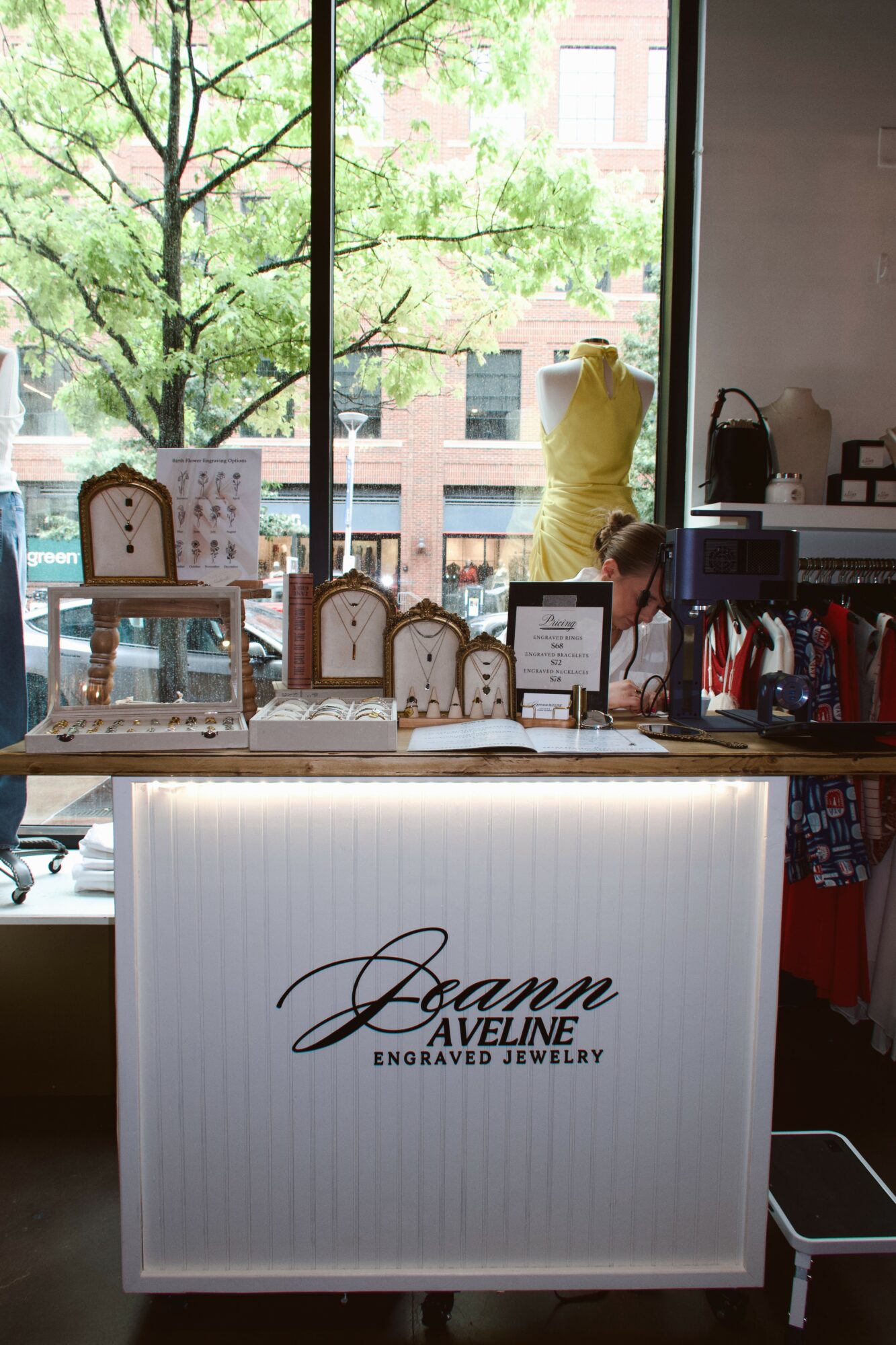 Jewelry display counter with a sign reading 'Seann Aveline Engraved Jewelry' inside a store, large window showing trees outside.