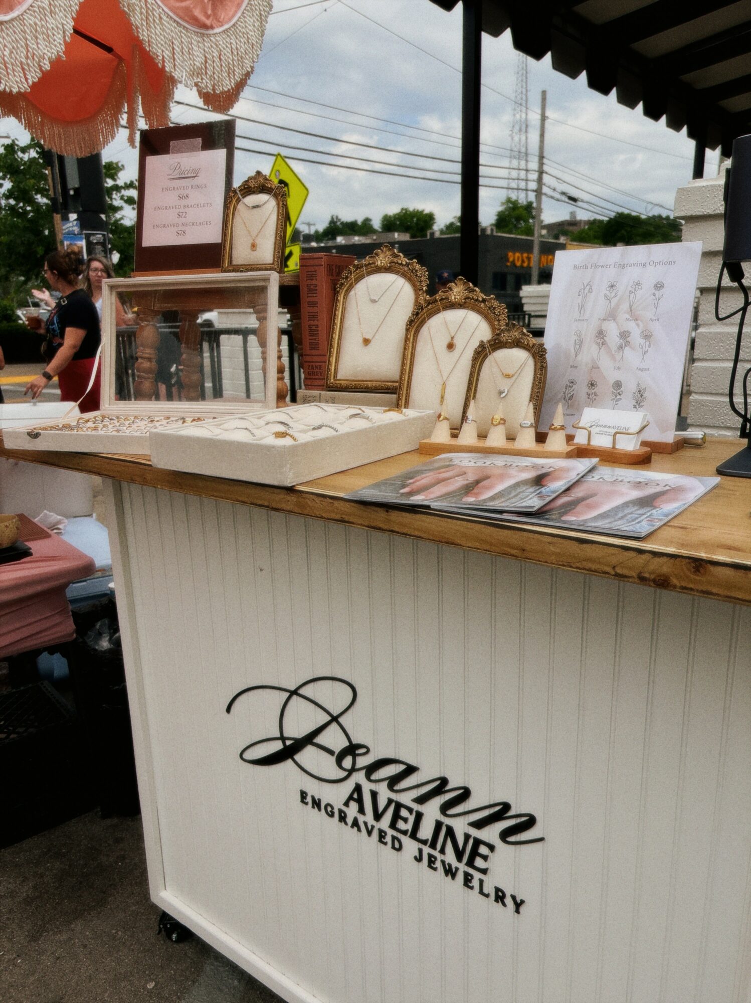 Jewelry display table with mirrors and jewelry, outdoor market setting, people in background, branded with 'Joann Avenue Engraved Jewelry'.