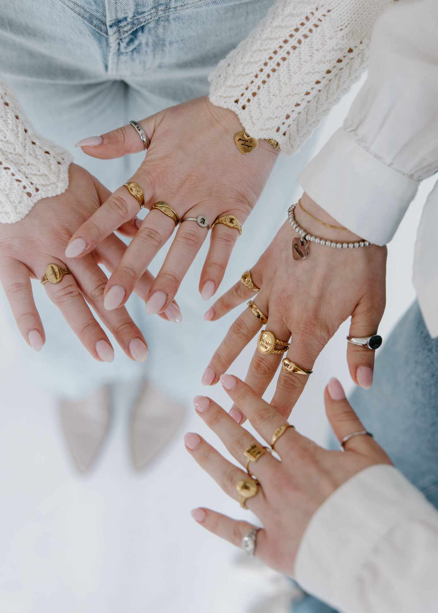 Multiple hands with rings and bracelets, wearing white and light-colored clothing, overlapping in a group gesture.
