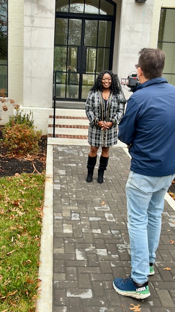 Woman with curly hair and glasses being interviewed outside a building, man with camera recording her, paved walkway, plants, and building facade.