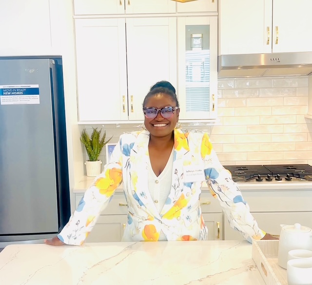 Smiling woman in a colorful blazer stands in a bright kitchen with white cabinets and a refrigerator.