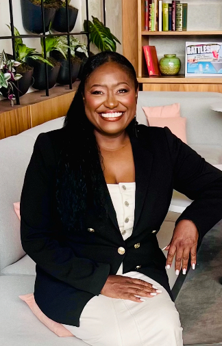 Smiling woman sitting on a sofa in a room with bookshelves and plants behind her.