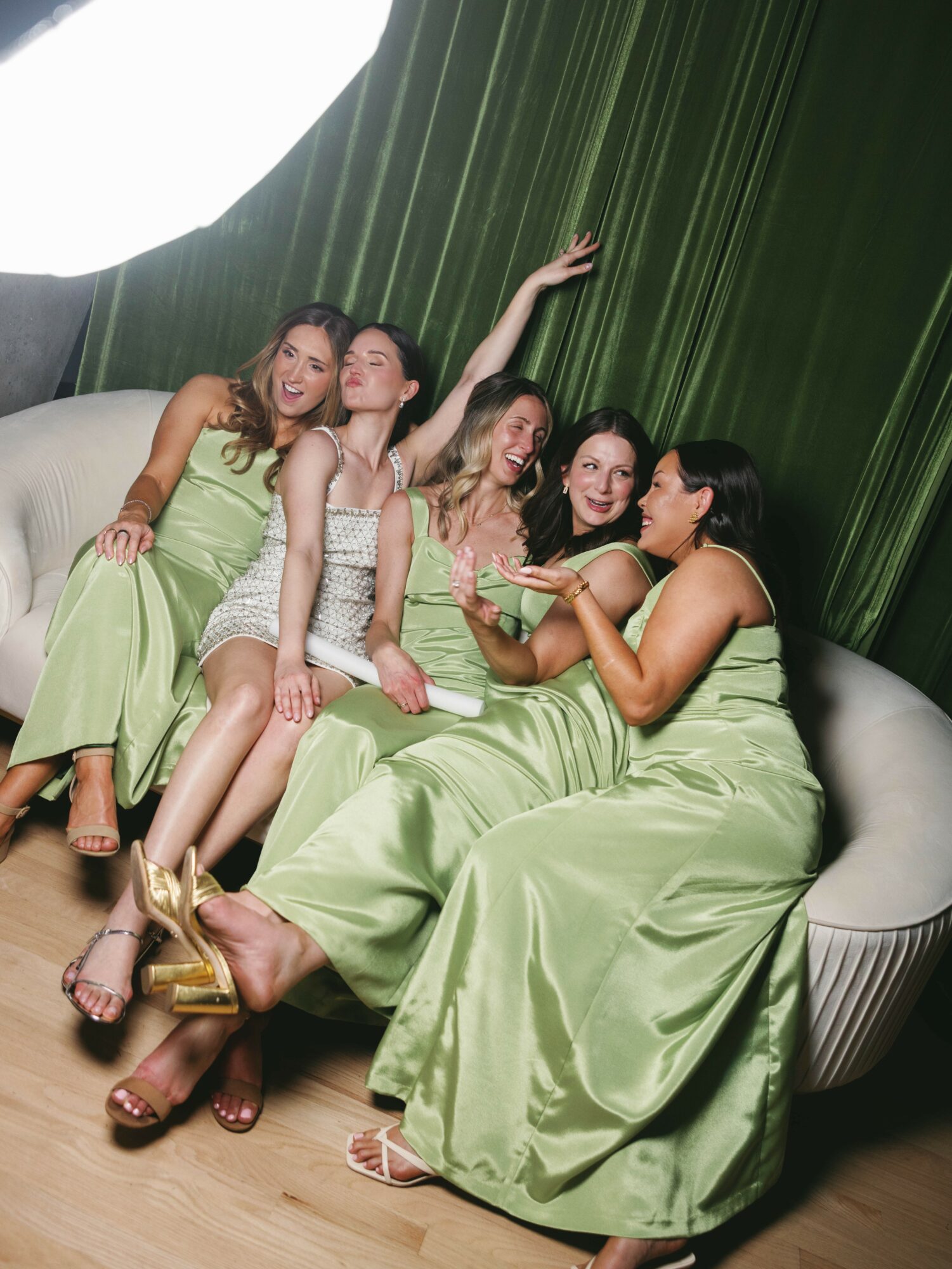 Five women in green dresses sitting on a curved sofa, with one woman holding a plate, in a room with green curtains.