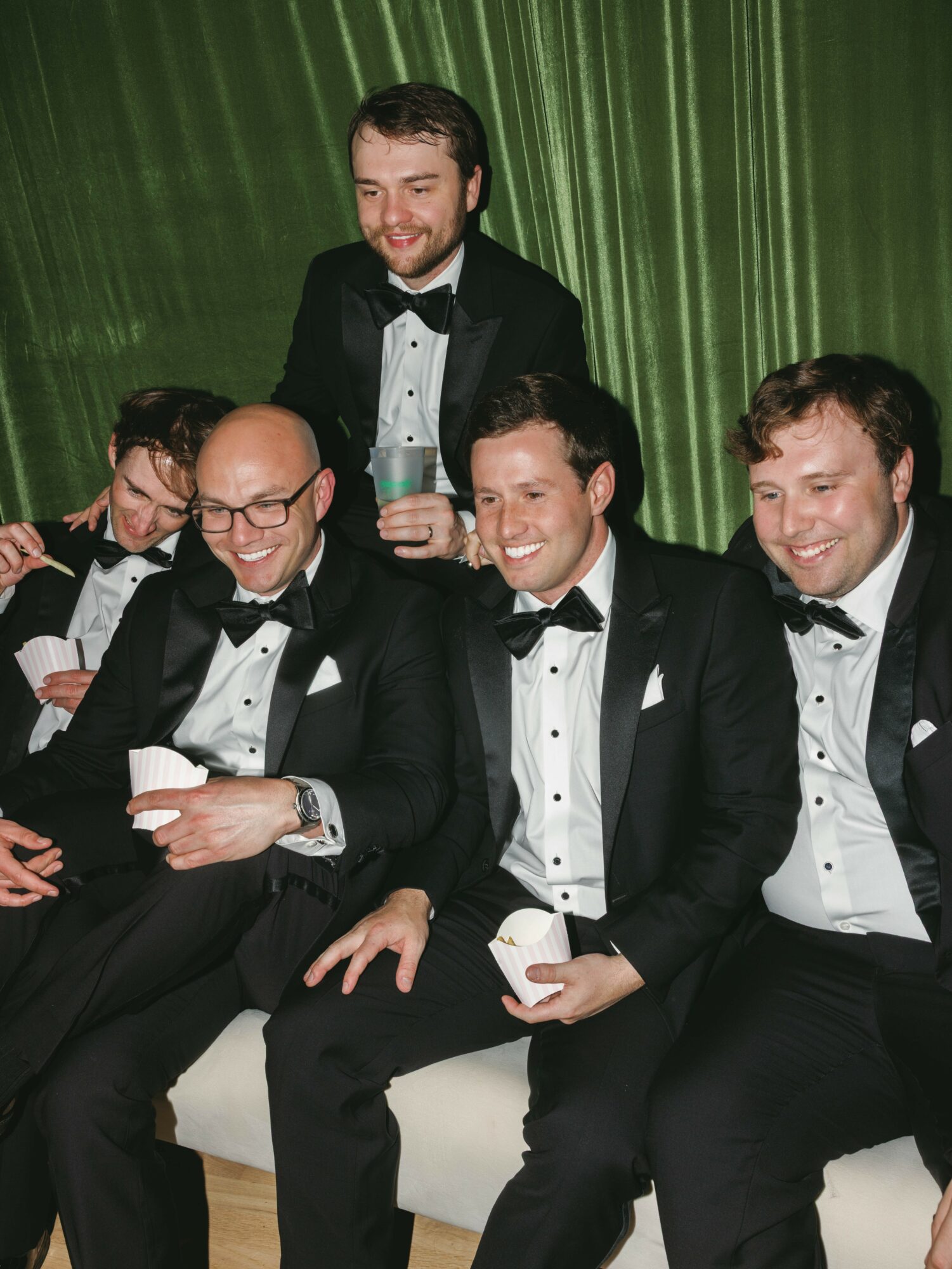 Group of five men in tuxedos sitting and standing, smiling, against a green curtain backdrop.