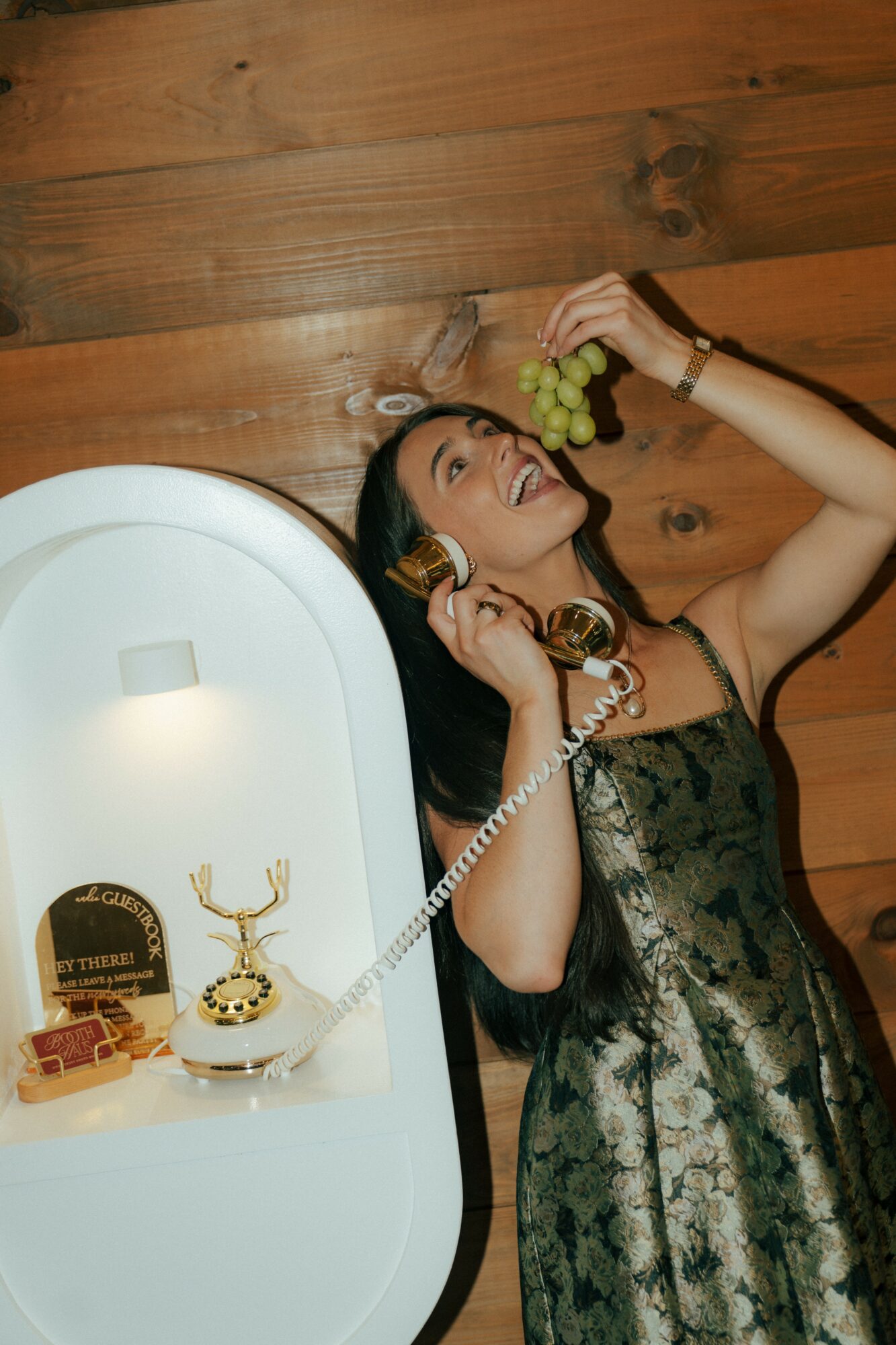 Woman with long dark hair talking on a vintage phone, holding a bunch of grapes, smiling, in front of a wooden wall.