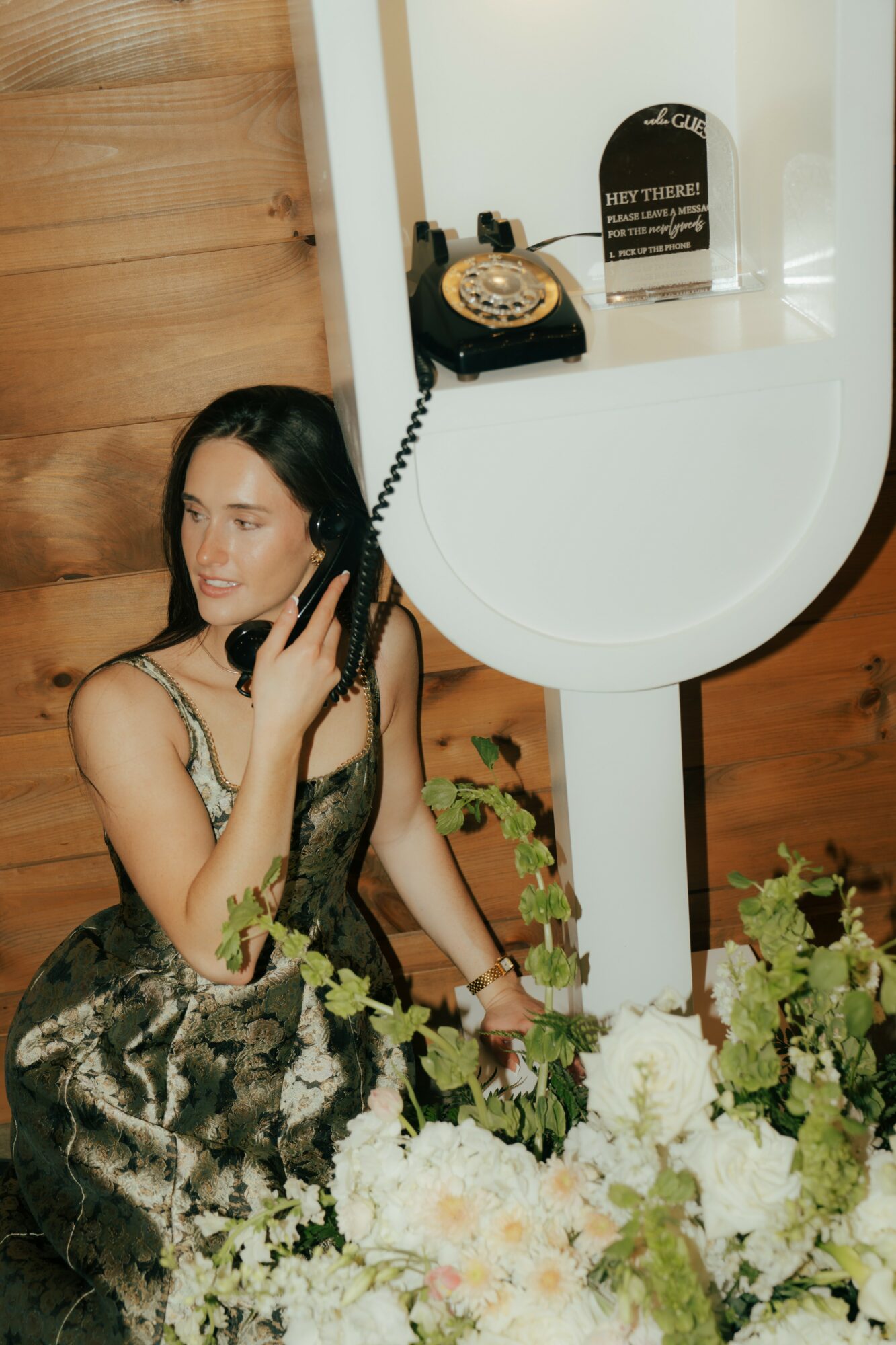 Woman in a dress talking on a vintage rotary phone, surrounded by flowers, with a wooden wall in the background.