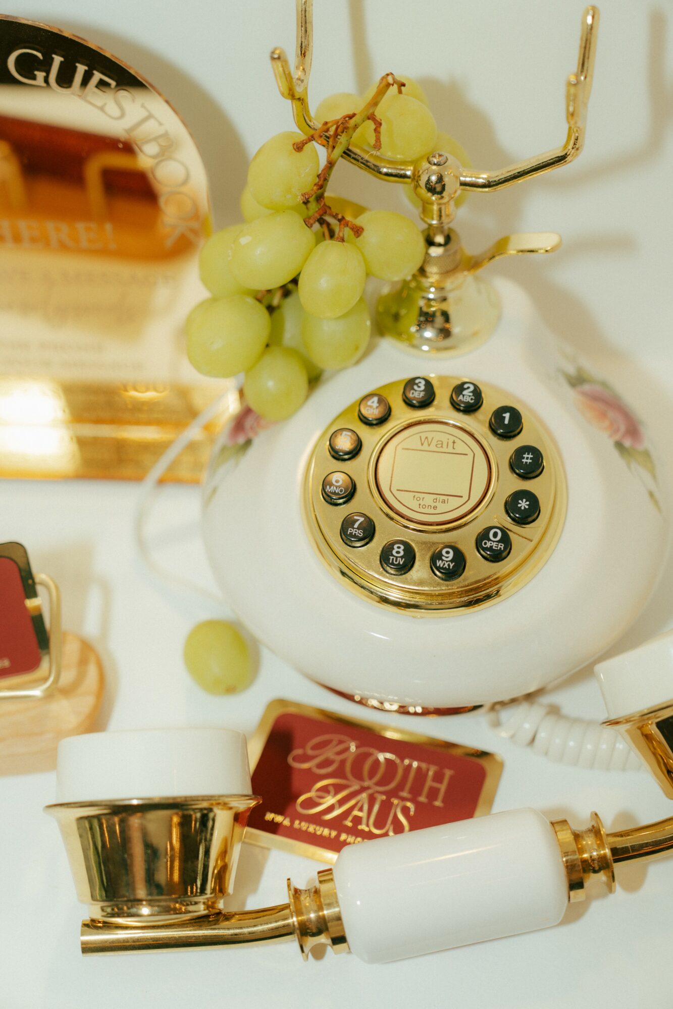 Vintage rotary telephone with grapes and jewelry on a table, surrounded by various objects, including a gold and white item and a red box.