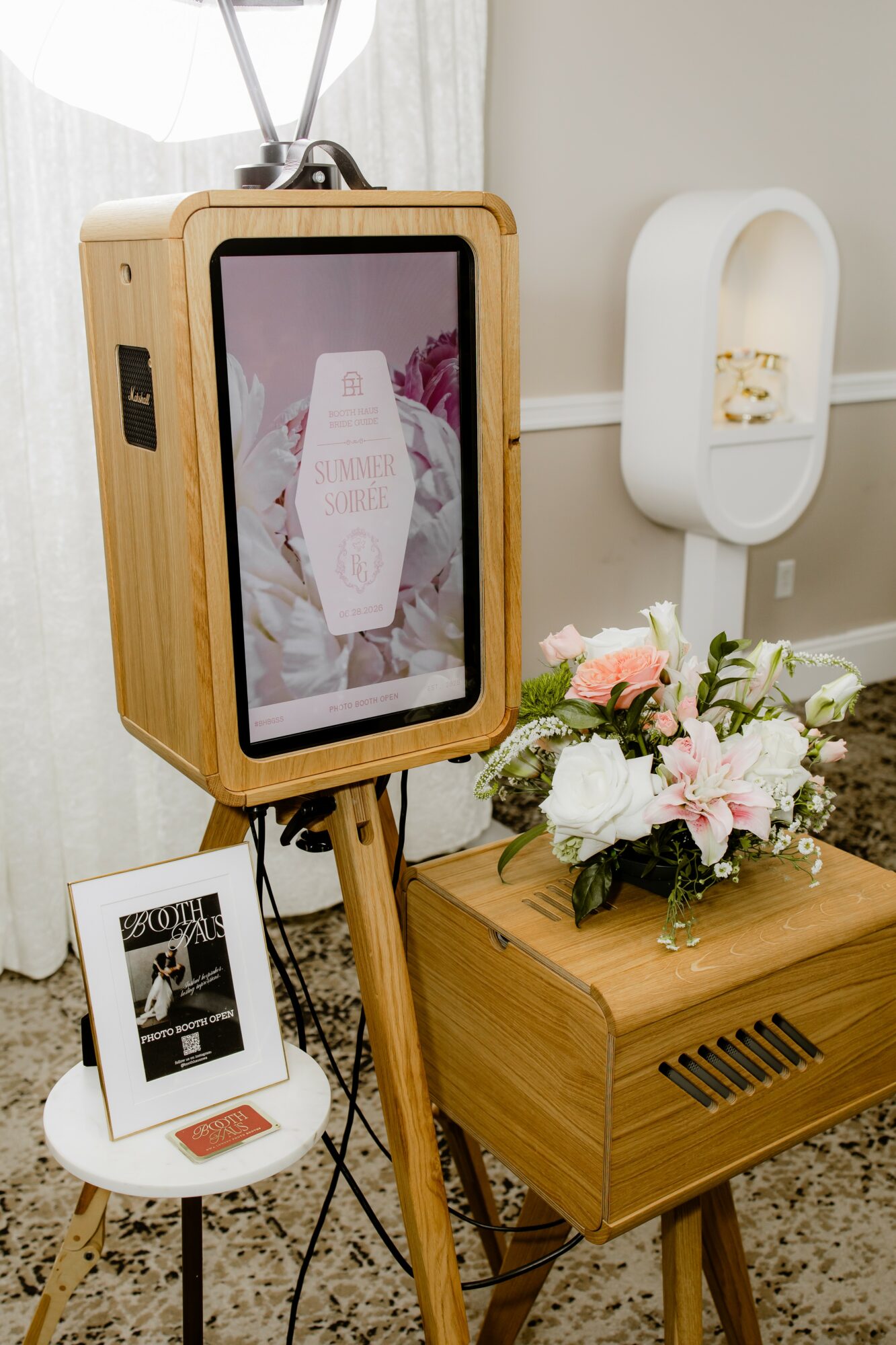 A wooden photo booth with a screen, a small white table with a photo, and a flower arrangement on a wooden table.