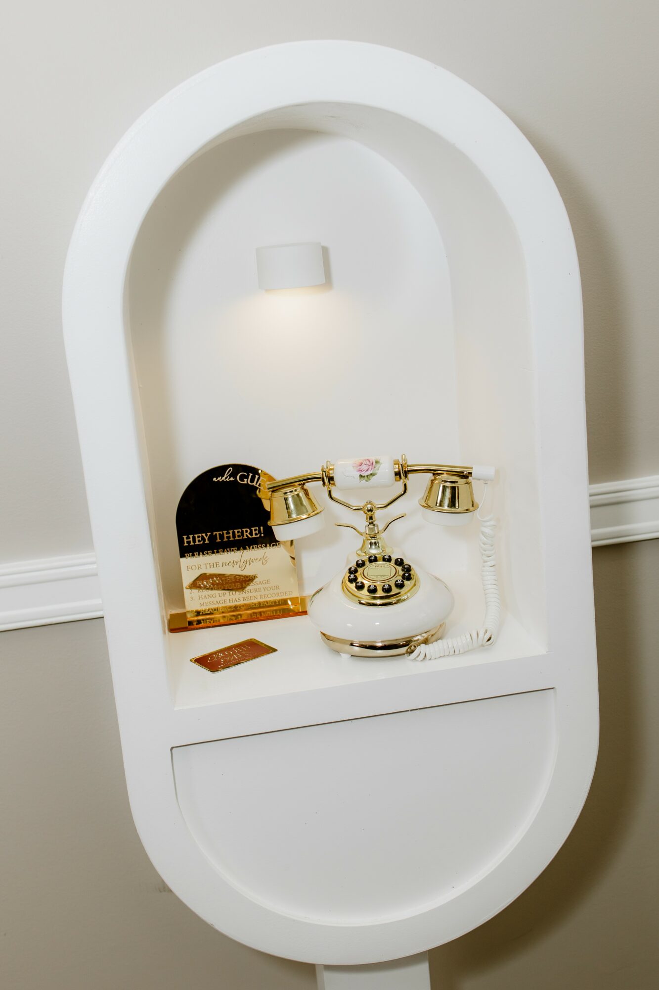 White oval display shelf with a gold and white telephone, a black and gold item, and small decorative objects, against a light wall.