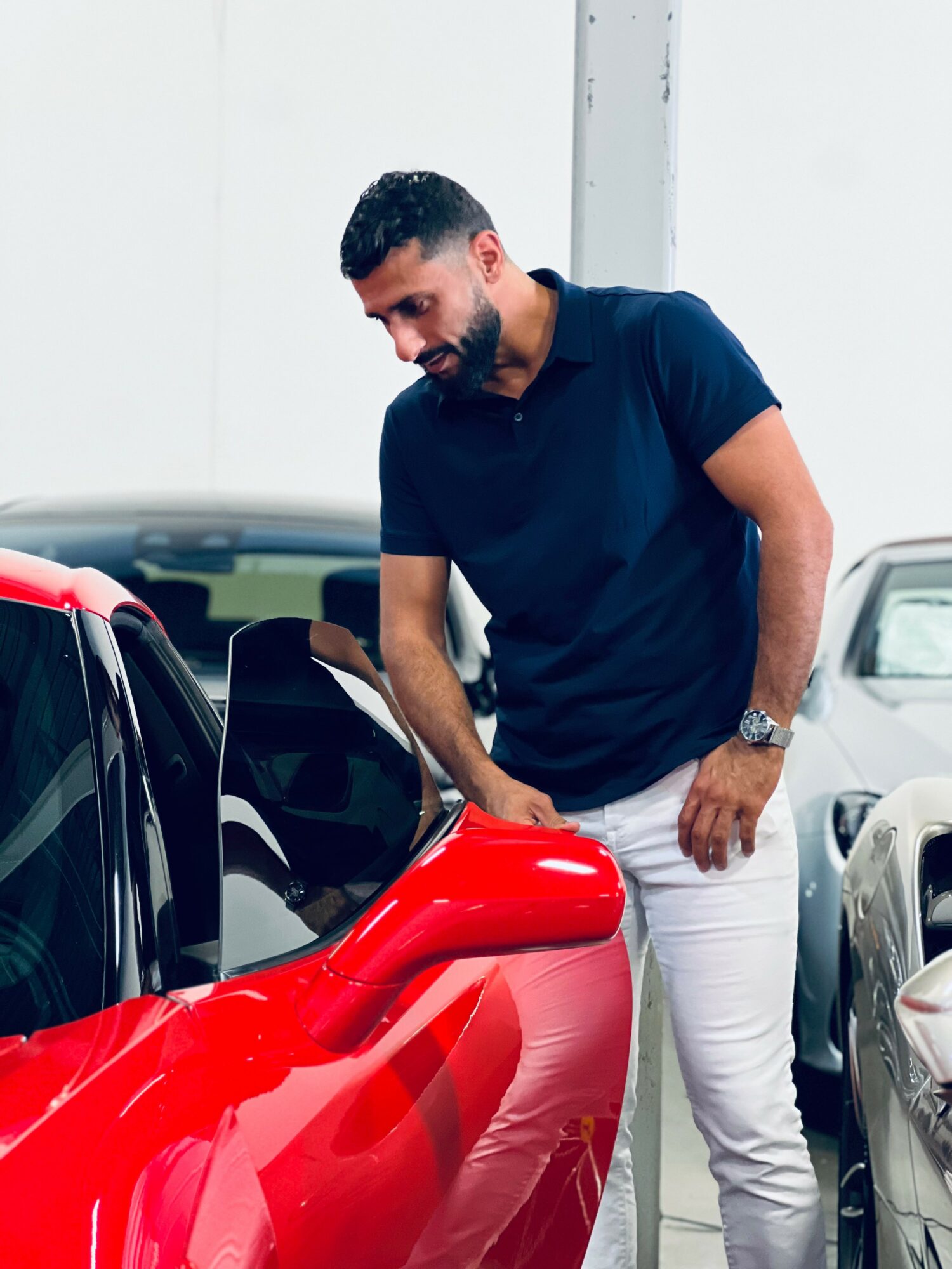 Man with dark hair and beard wearing a navy blue shirt and white pants, smiling while looking at a red car.