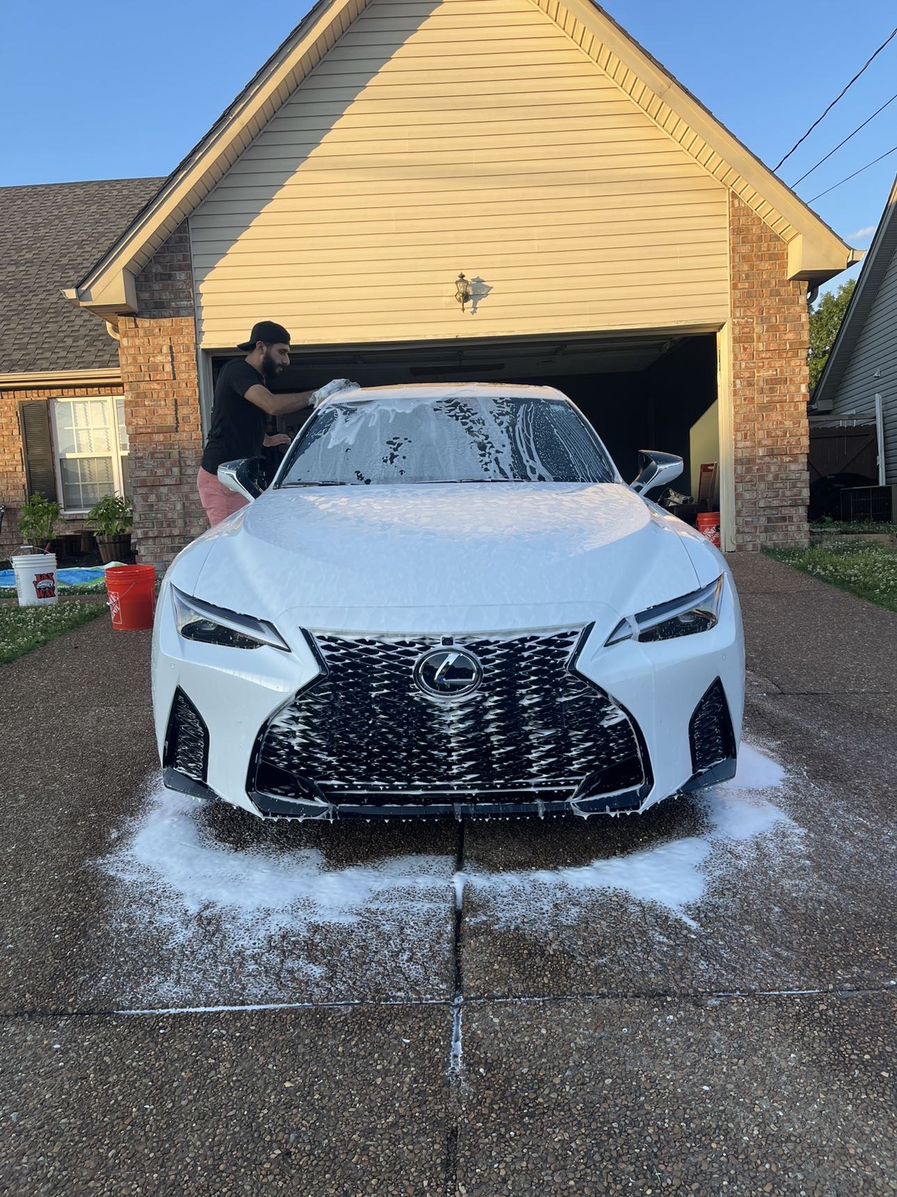 Person washing a white Lexus car in driveway with house in background.