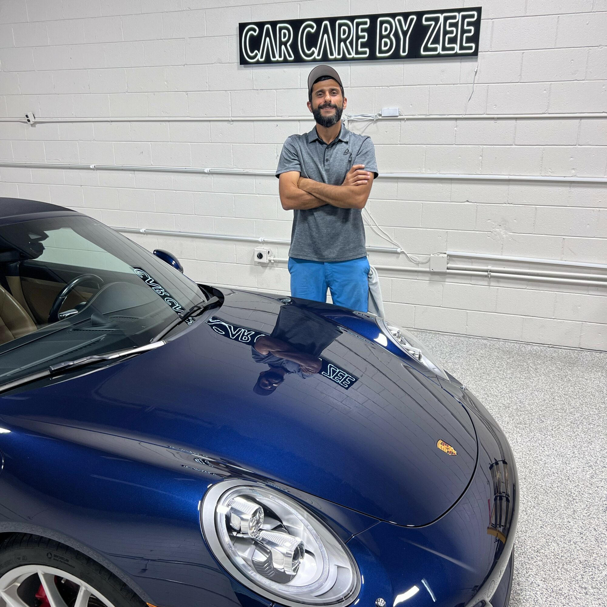 Man with beard and crossed arms standing next to a blue car in front of a white wall with a sign that reads 'CAR CARE BY ZEE'.