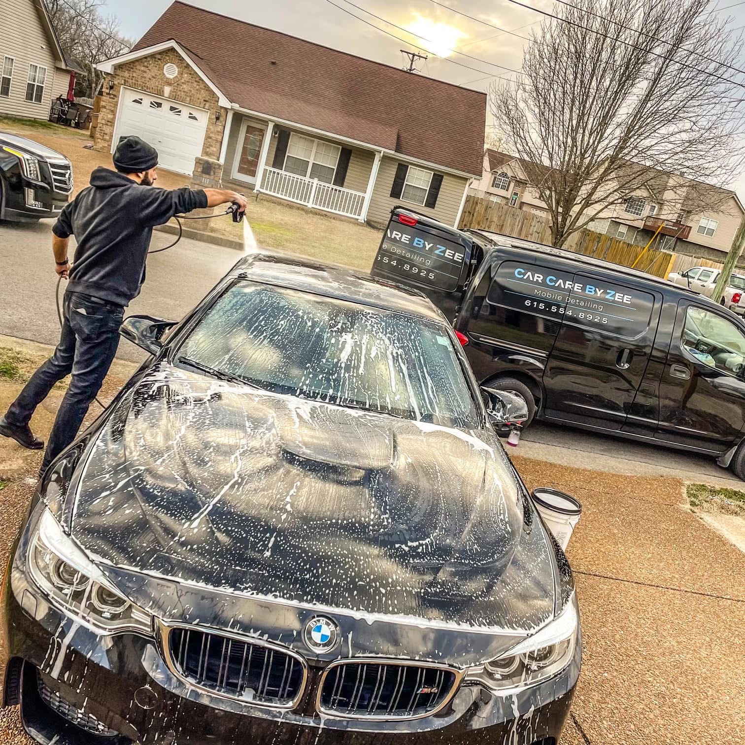 Person washing a black BMW car with a hose on a residential street, houses and trees in background.