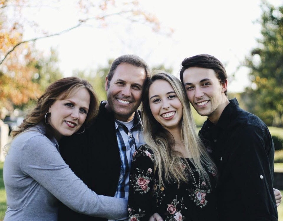 Four people smiling outdoors, standing close together, with trees and sky in the background.