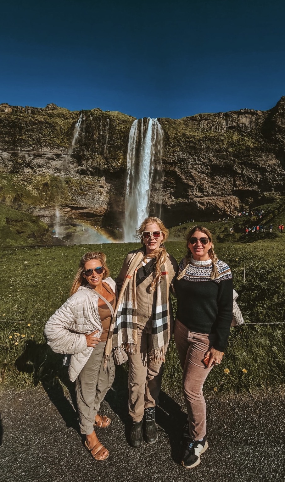 Three women stand in front of a waterfall with a rocky cliff and blue sky, smiling outdoors.