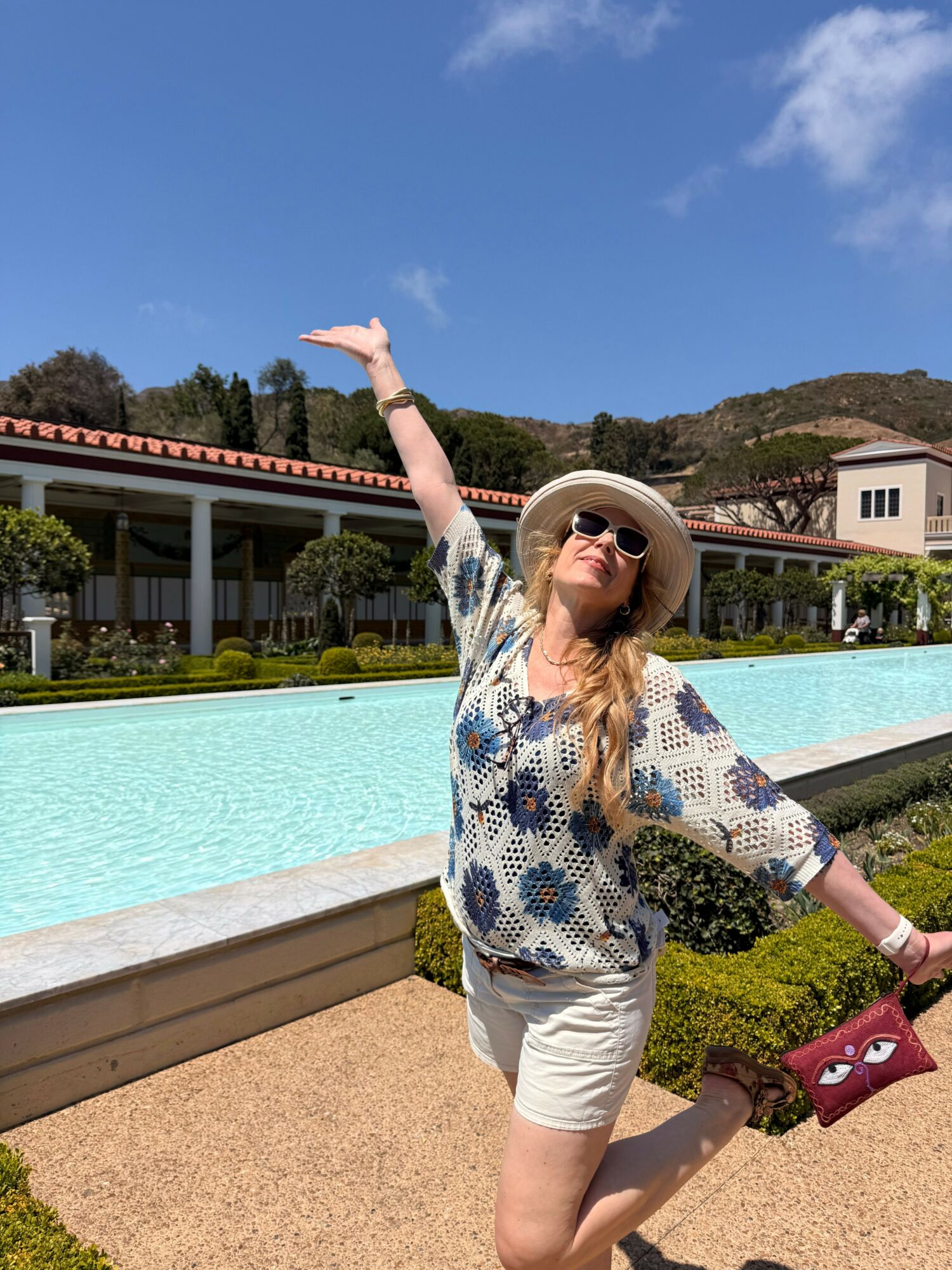 Woman in hat and sunglasses reaches out by poolside on sunny day, with buildings and trees in background.