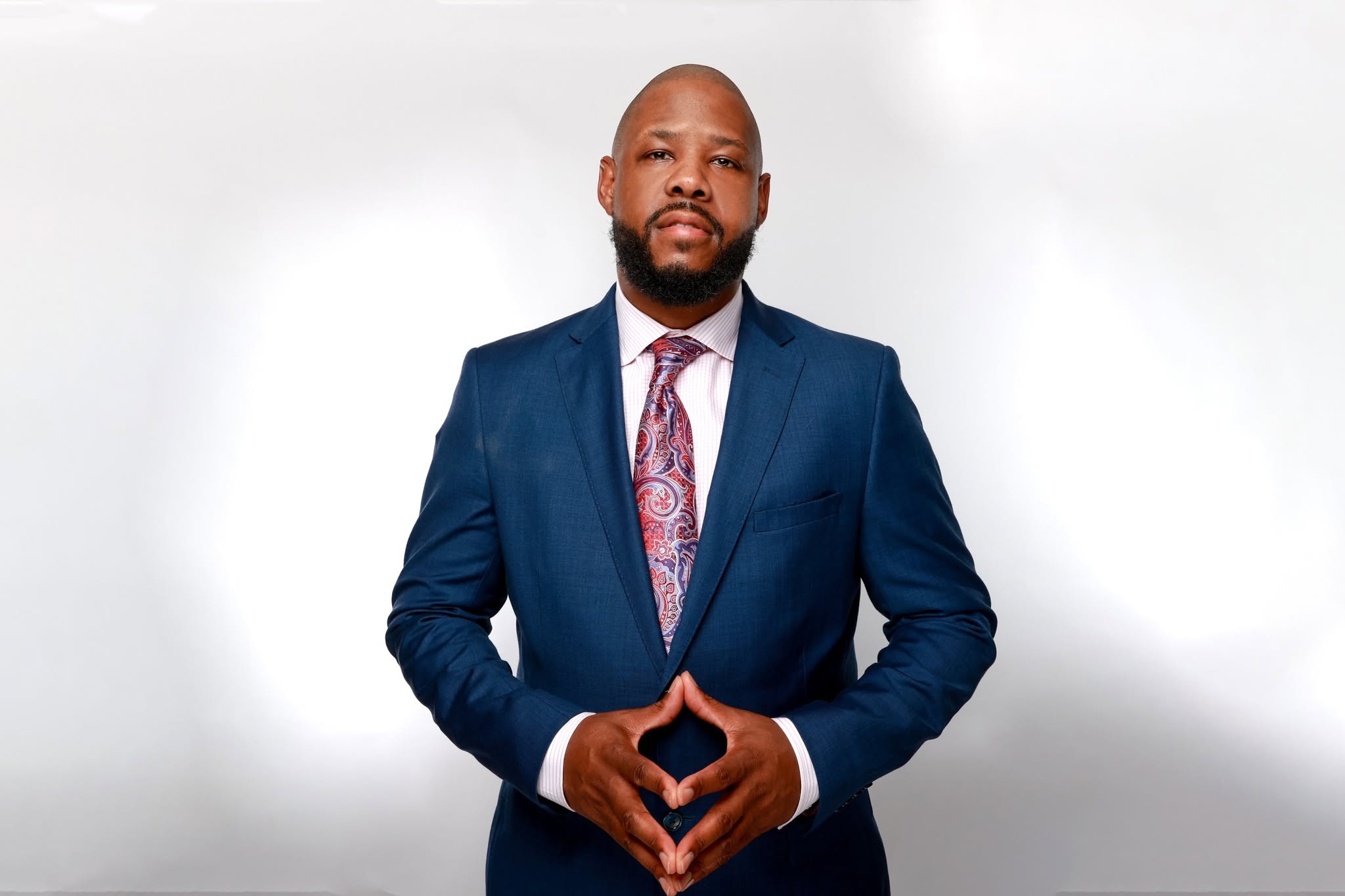 Man in a blue suit and tie standing with hands clasped in front, neutral expression, plain white background.