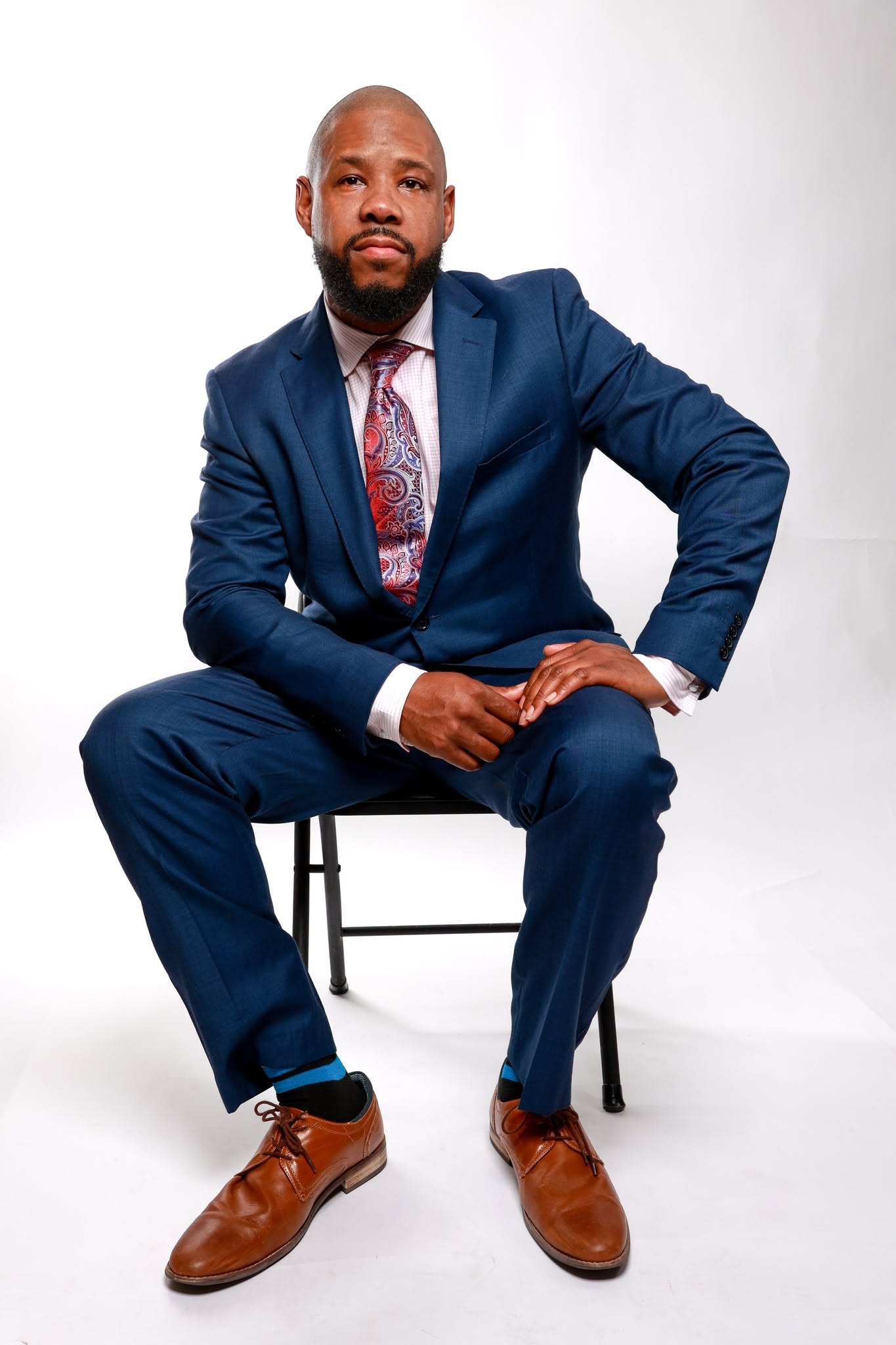 Man in a blue suit sitting on a chair, white background, looking at the camera, with a serious expression.