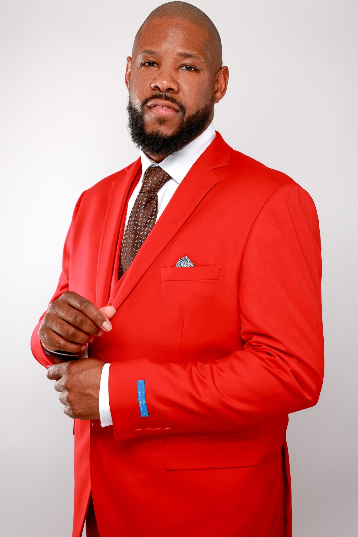 Man in red suit adjusting his jacket, wearing a white shirt and brown tie, with a beard and bald head.