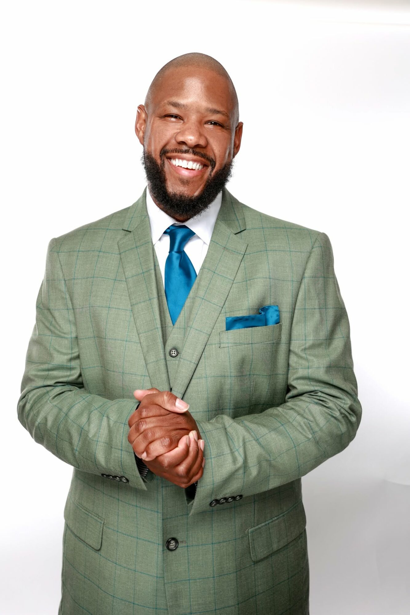 Man in a green suit with a blue tie and pocket square, smiling with hands clasped, against a white background.