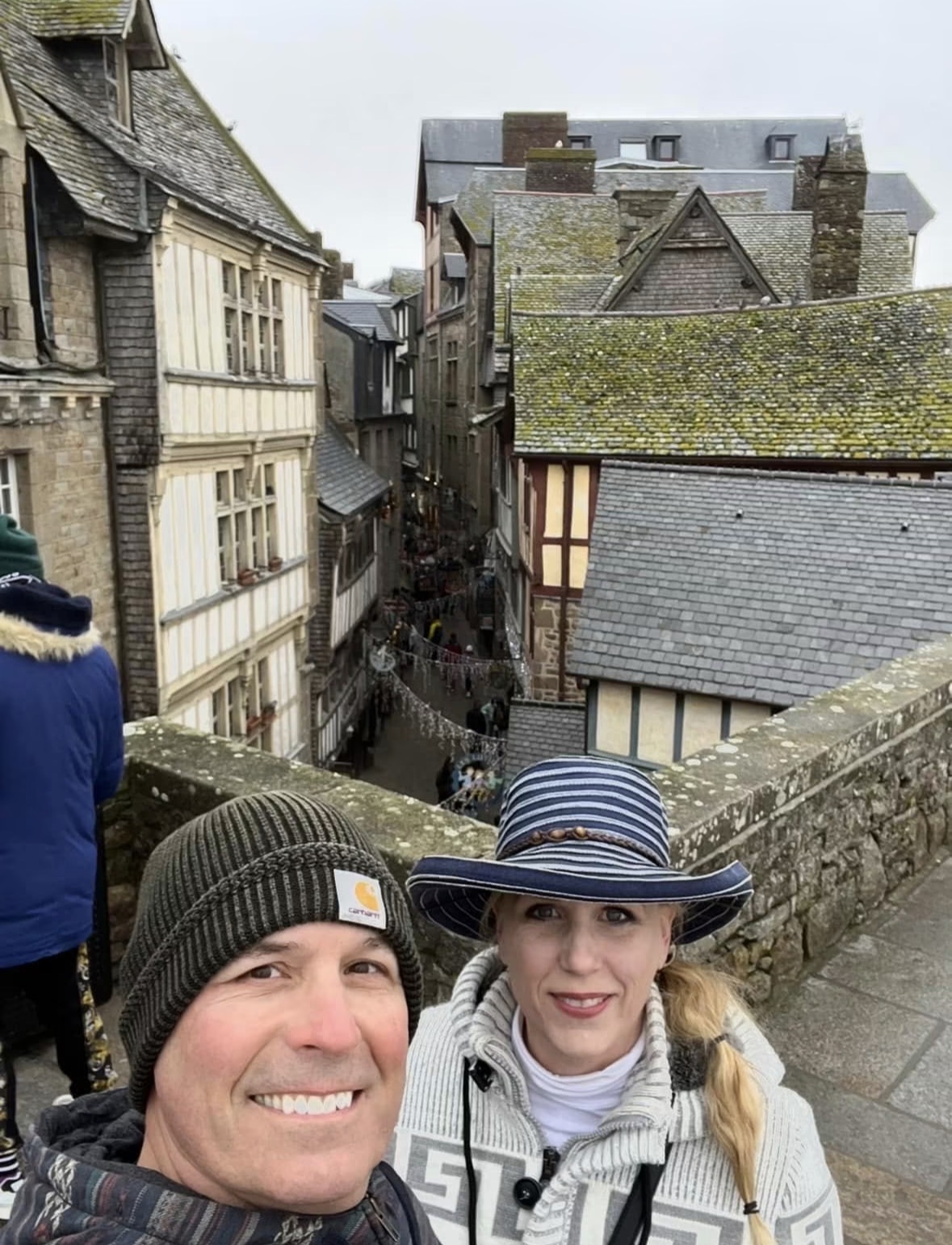 Two people taking a selfie on a stone bridge overlooking a narrow street with historic buildings, some with moss-covered roofs.