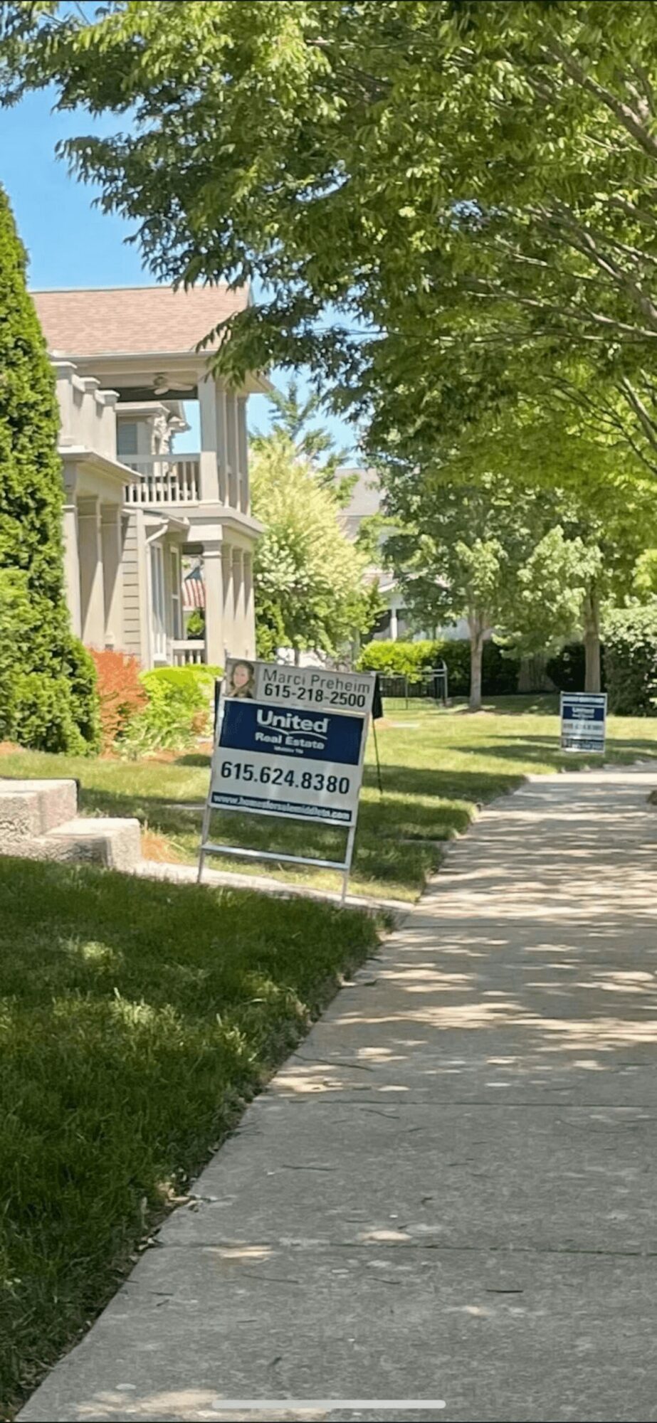 Sidewalk with trees and signs in front of residential buildings, sunny day, green foliage, blue sky.