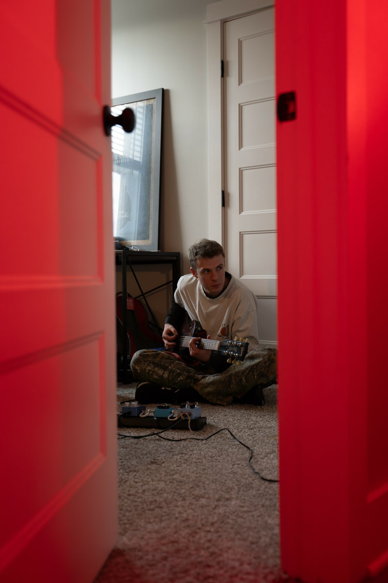 Person sitting on carpeted floor playing guitar in a room, viewed through a partially open door, with a mirror and door in background.