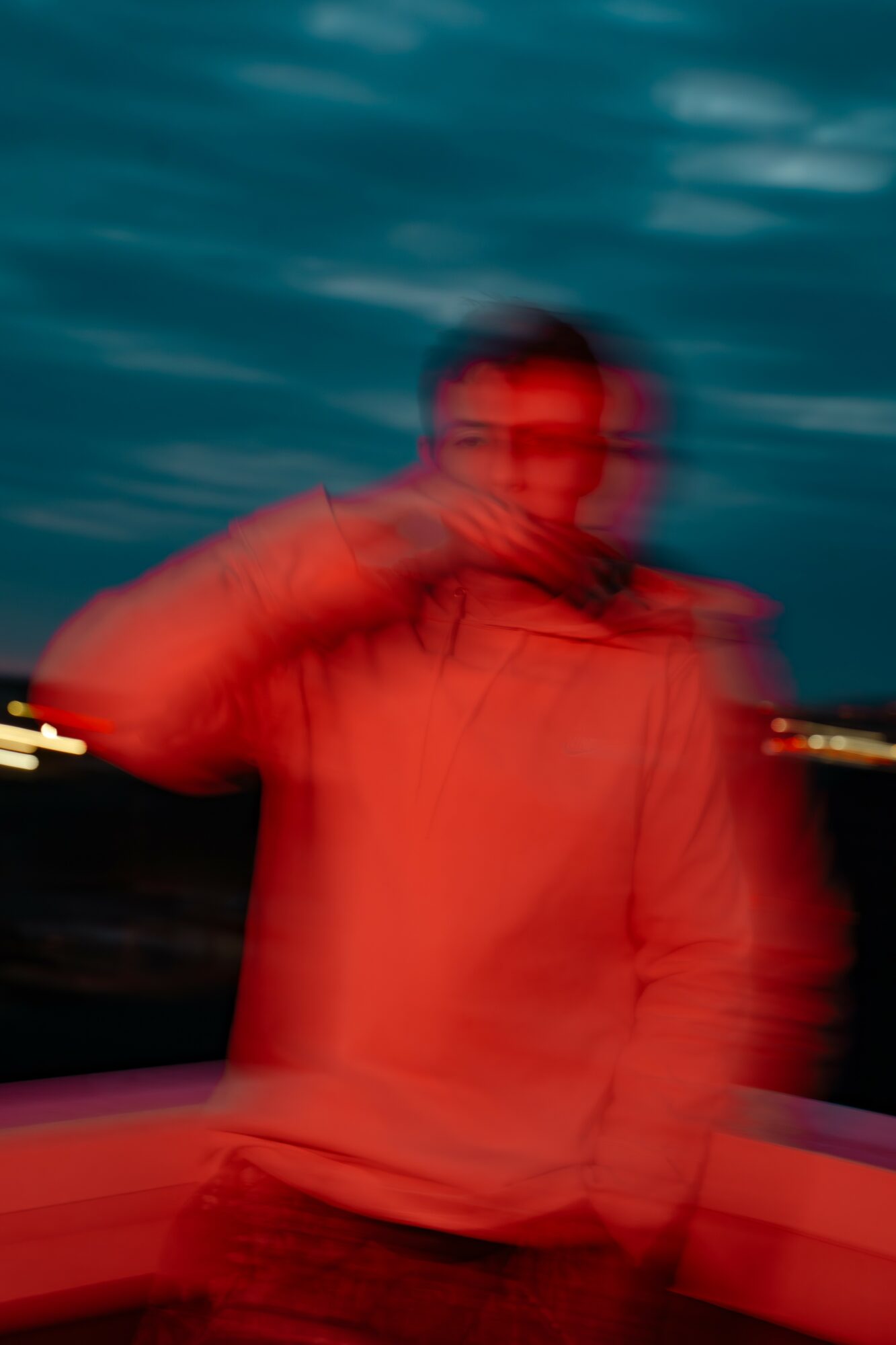 Person standing outdoors near water, blurred due to motion, wearing a jacket, with city lights in the background.