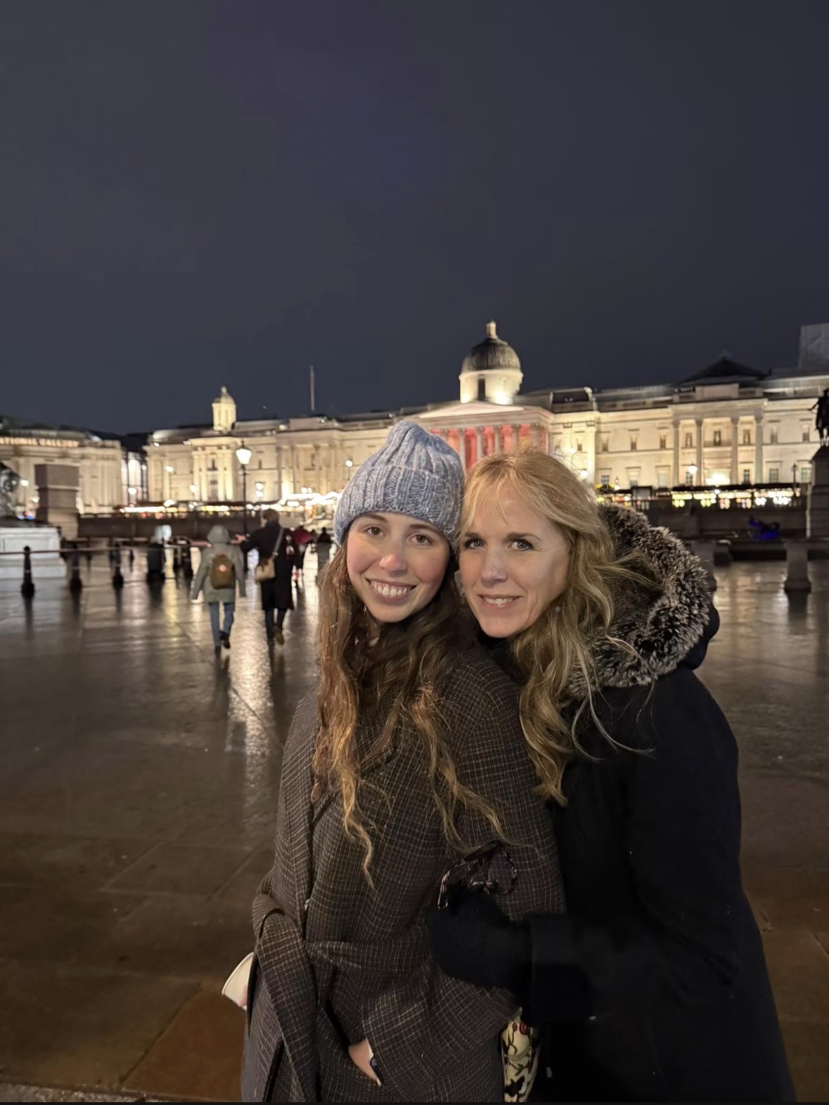 Two women smiling in front of a lit historic building at night, with a dark sky and wet ground.