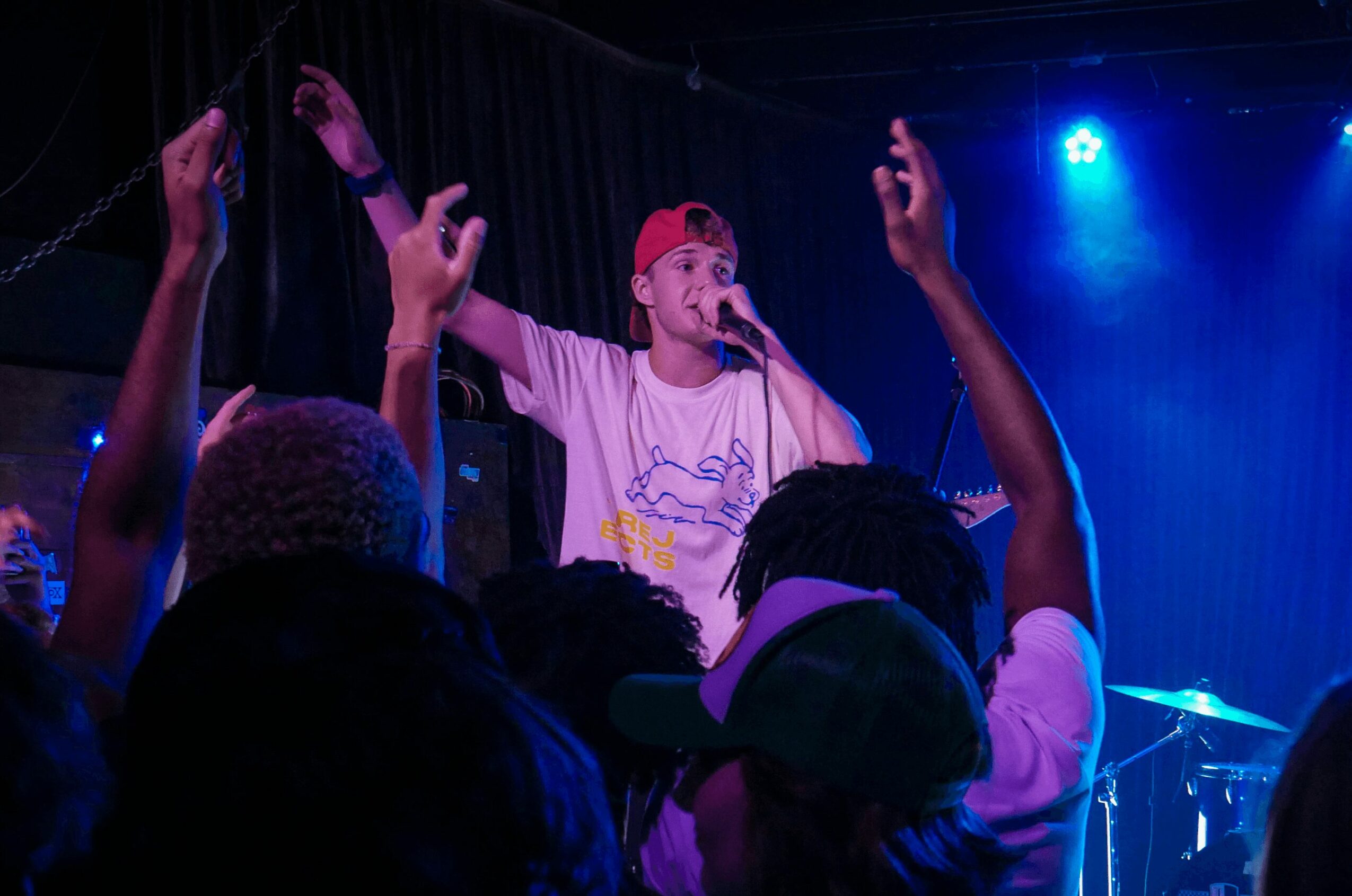 Performer on stage with arms raised, audience in foreground, blue stage lighting, musical instruments visible.