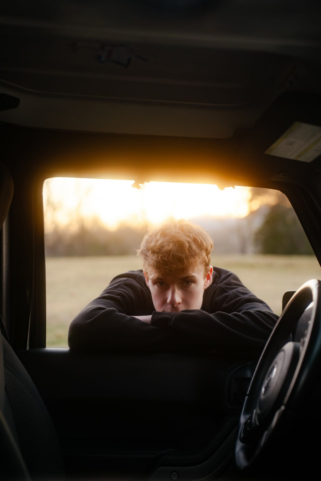 Person with curly hair leaning on car window, looking outside during sunset, with a landscape background.