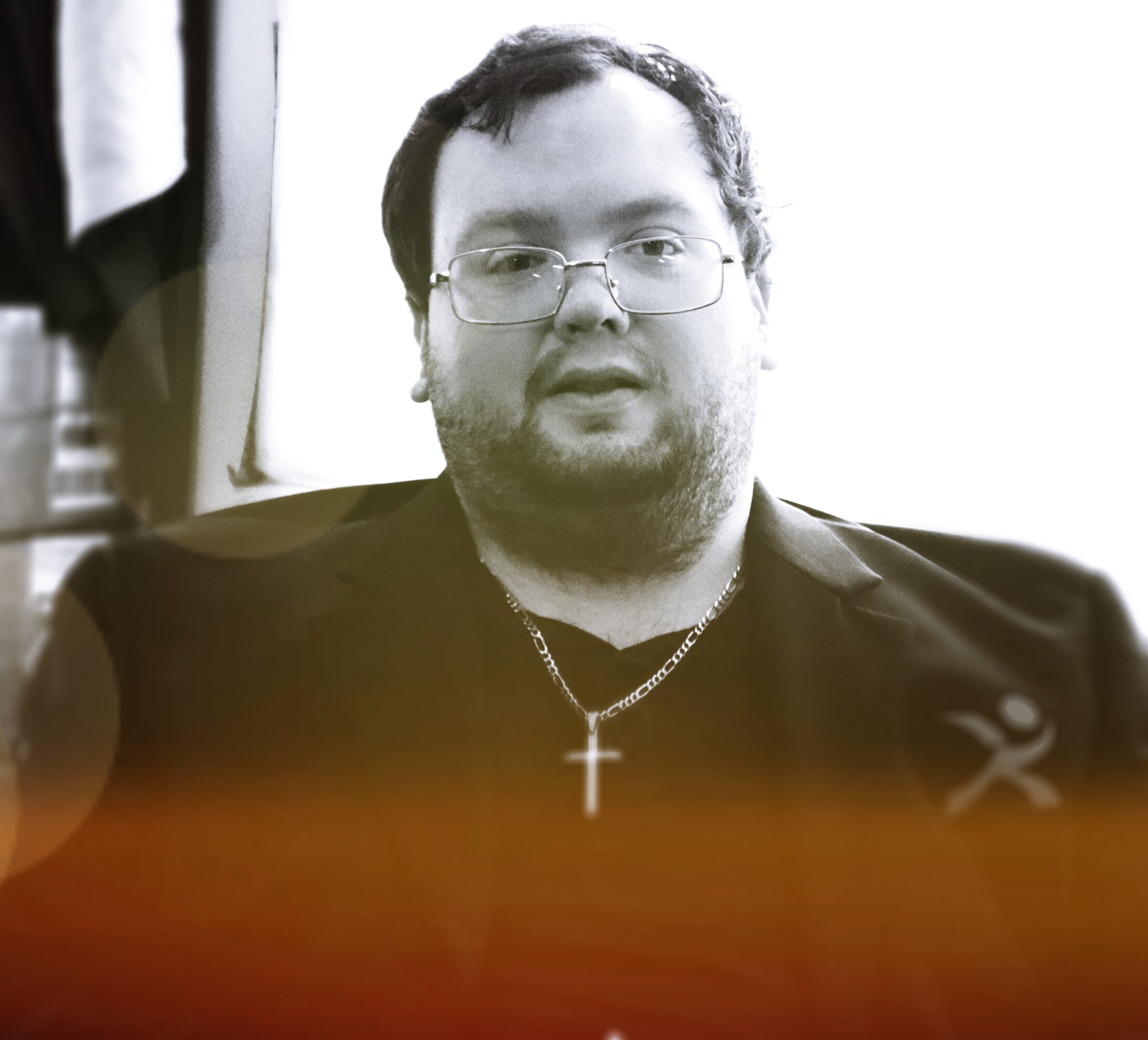 Man with glasses, beard, and cross necklace, wearing a dark jacket, sitting indoors with a blurred background.