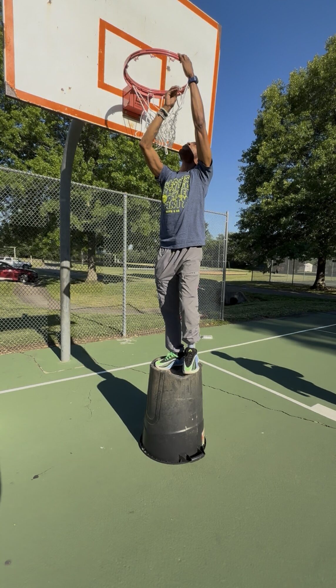 Person standing on a black platform, reaching up to hang a basketball net on an outdoor court with trees and a fence in background.
