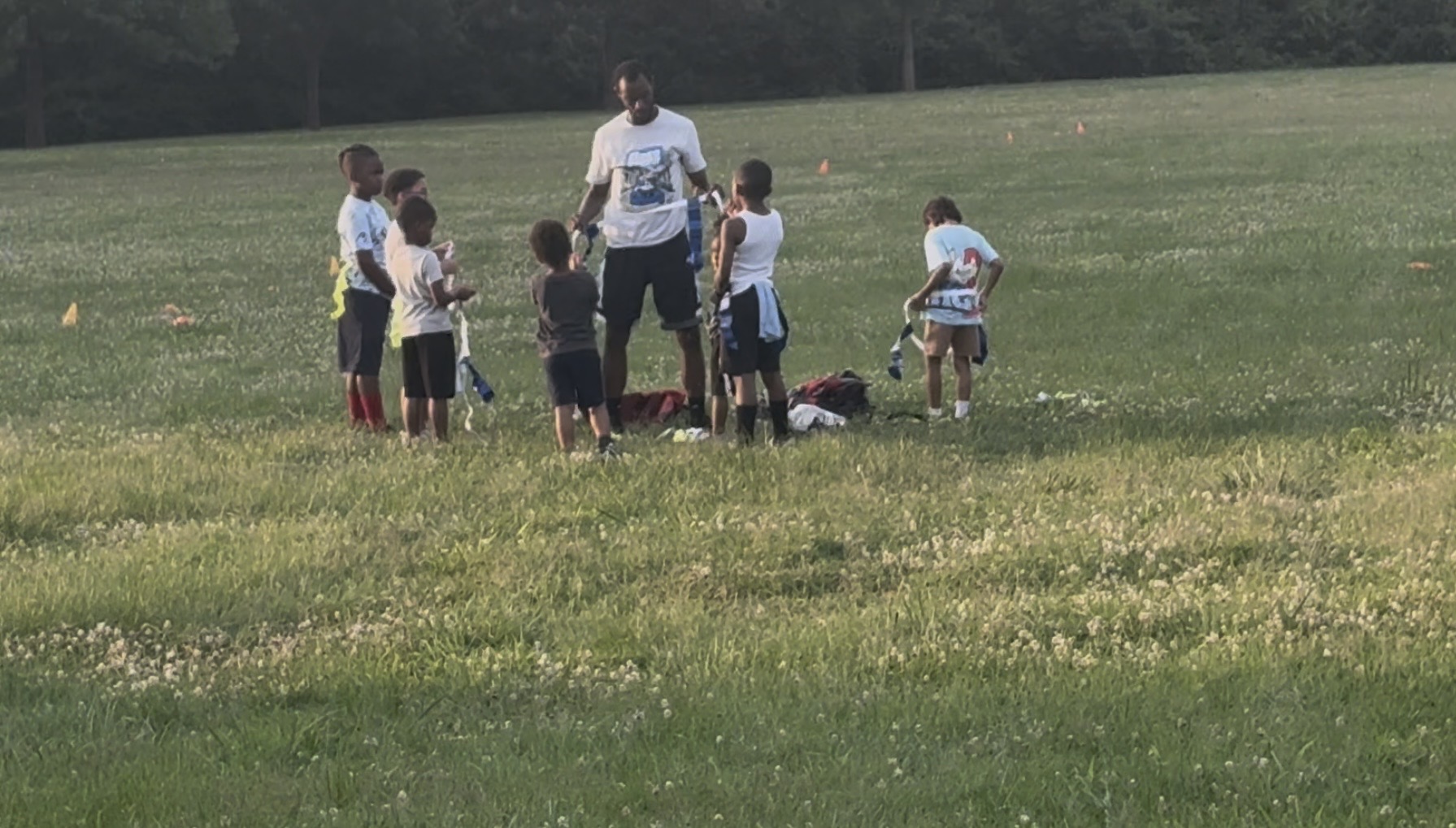 Group of children and an adult standing on a grassy field, some children sitting or standing, in a park-like setting.