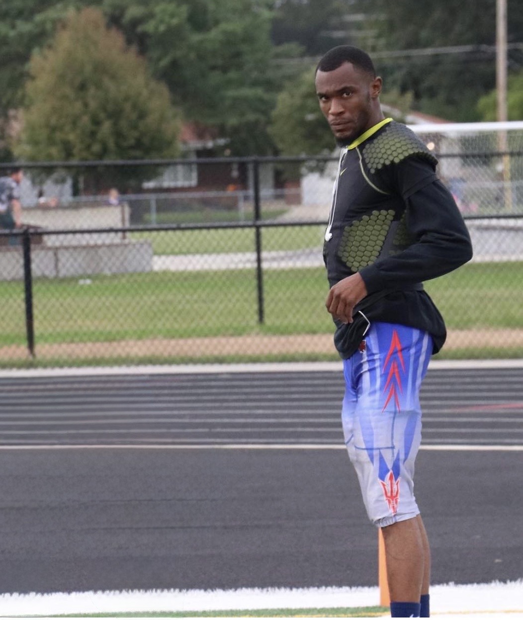 Young man standing on a track field with a fence and trees in the background, wearing a black and green jacket and blue shorts.