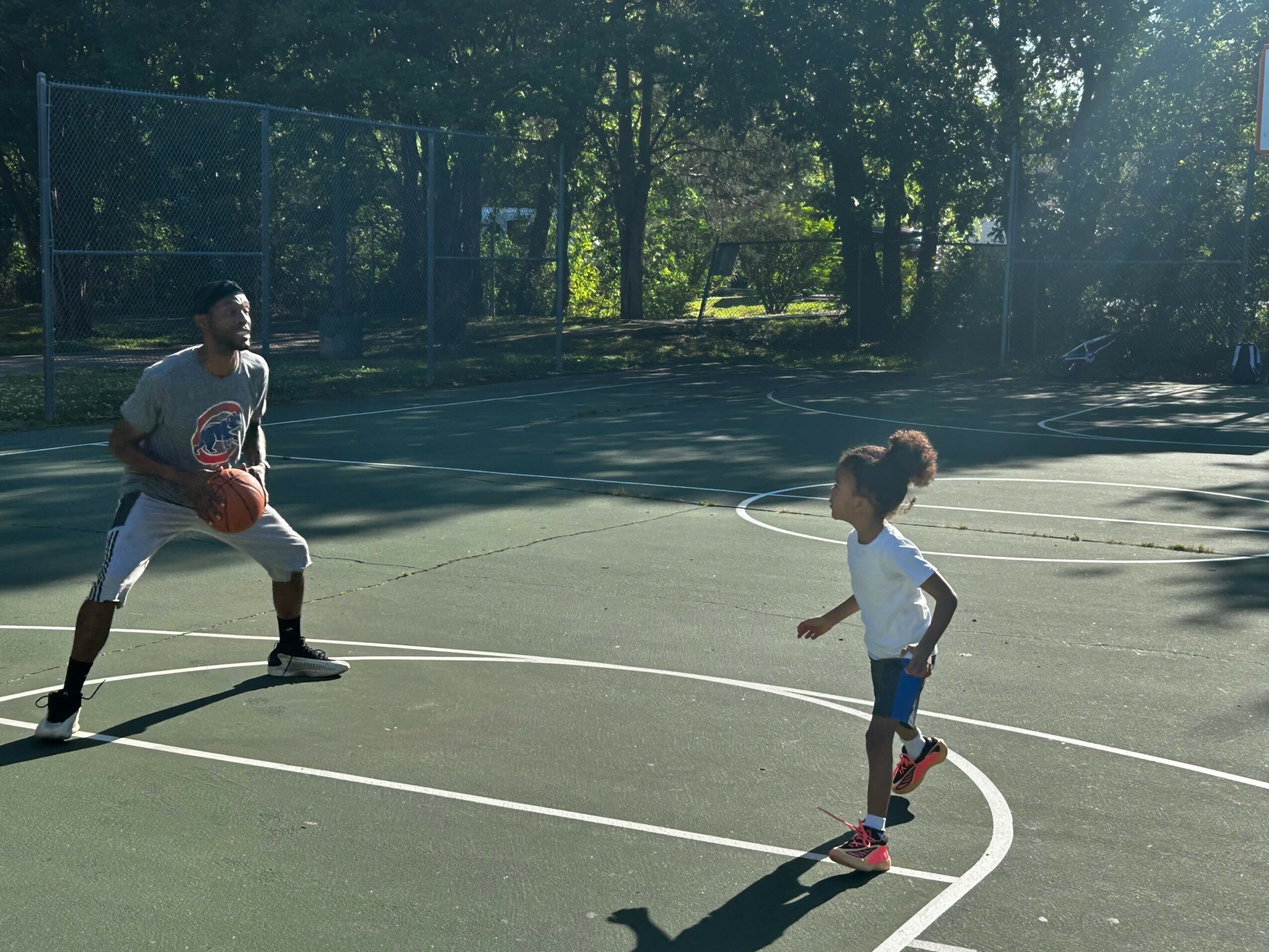 Two people playing basketball on an outdoor court with trees in the background.