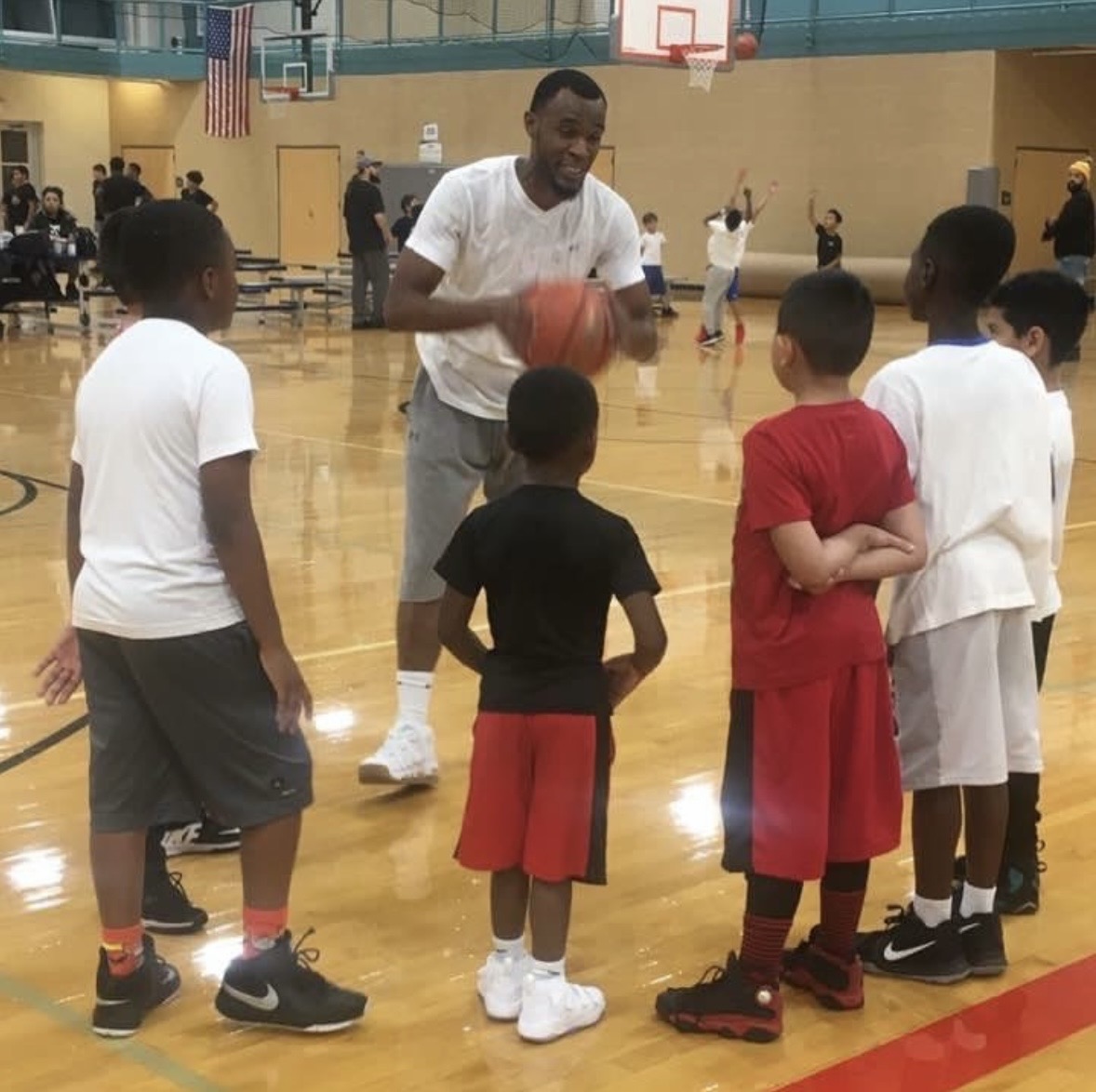A man in a white shirt holding a basketball talks to children in a gymnasium, with others practicing in the background.