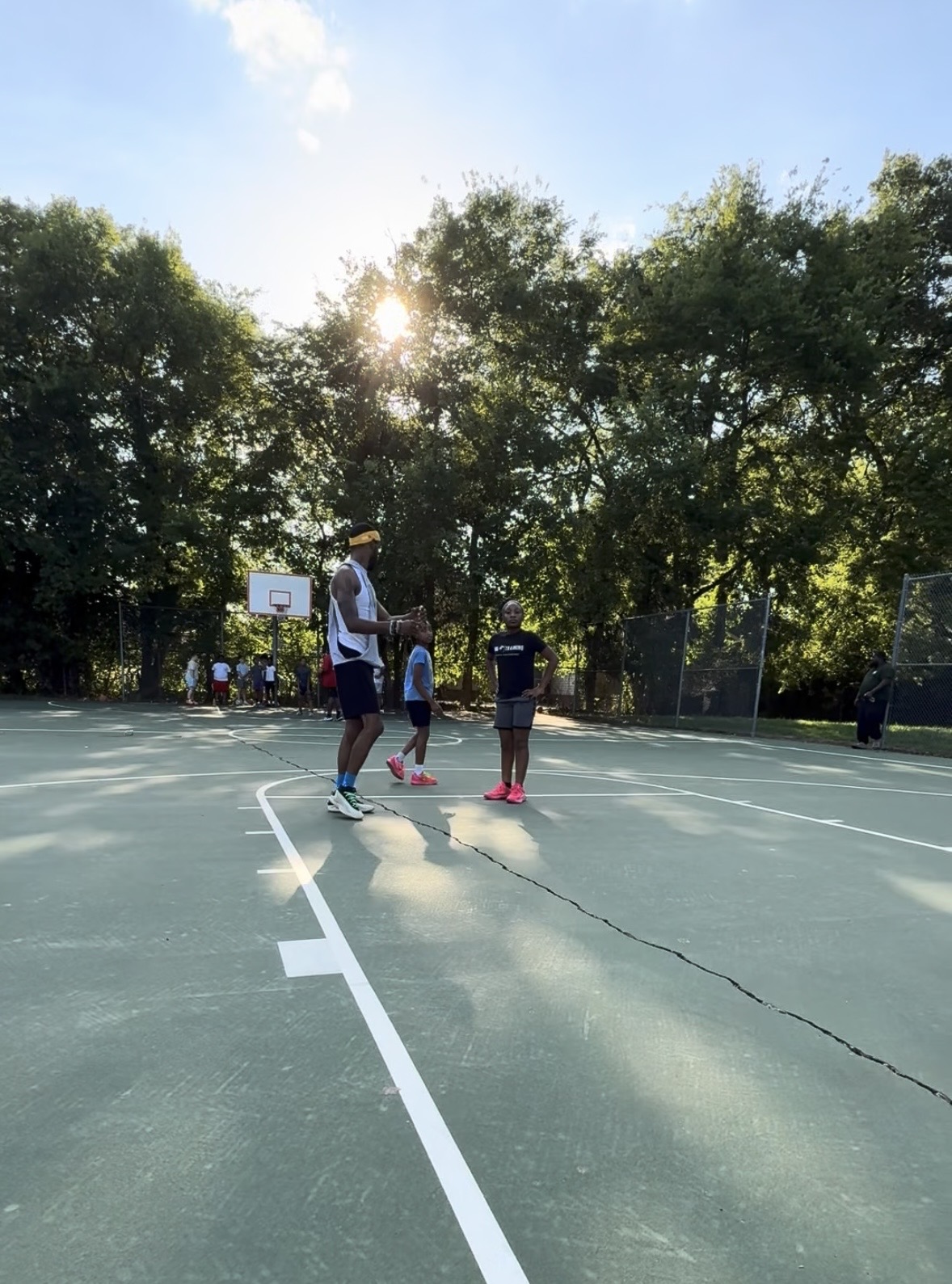 People playing basketball on an outdoor court with trees and sunlight in the background.