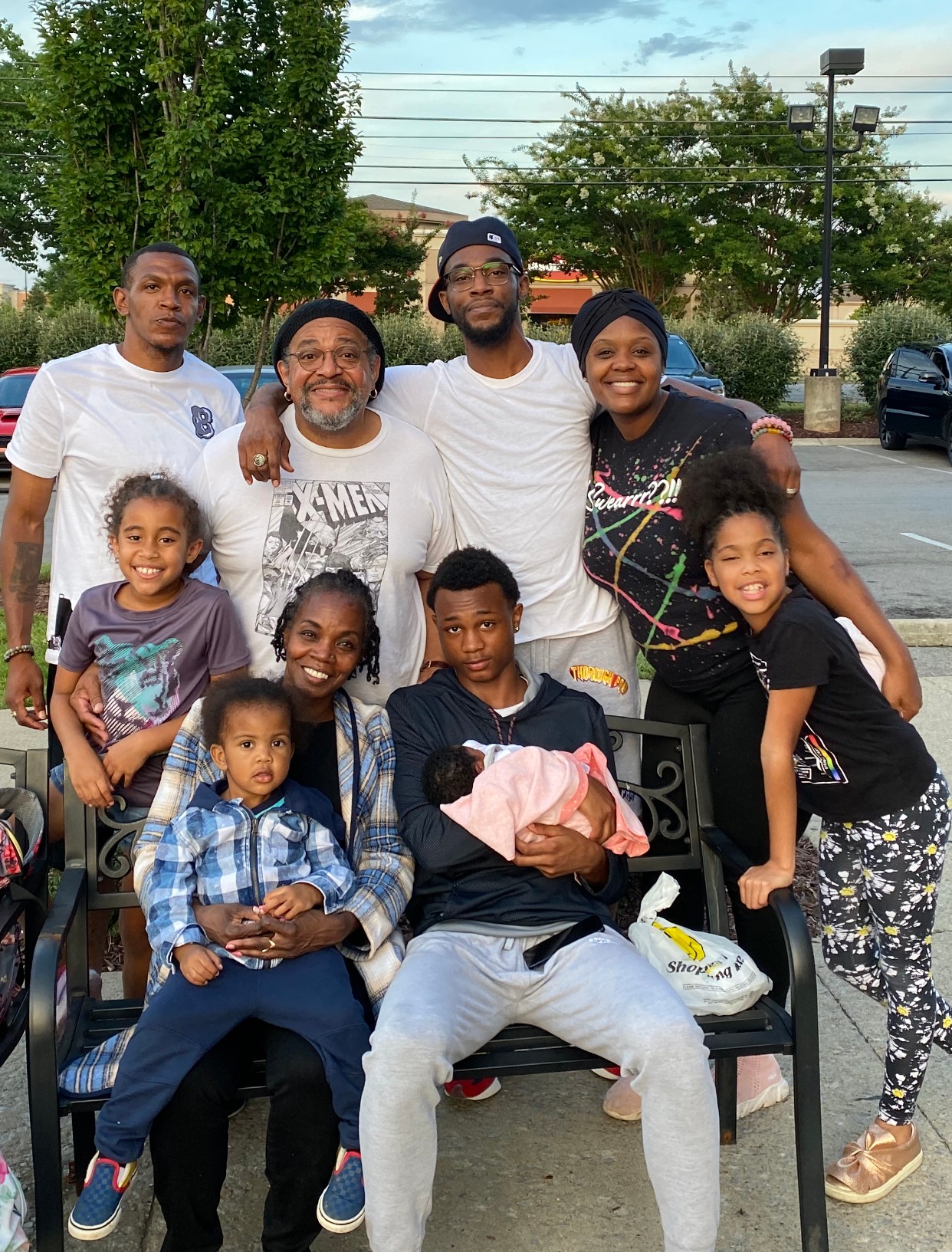 Group of nine people, including children and adults, outdoors in a parking lot with trees and cars in background.