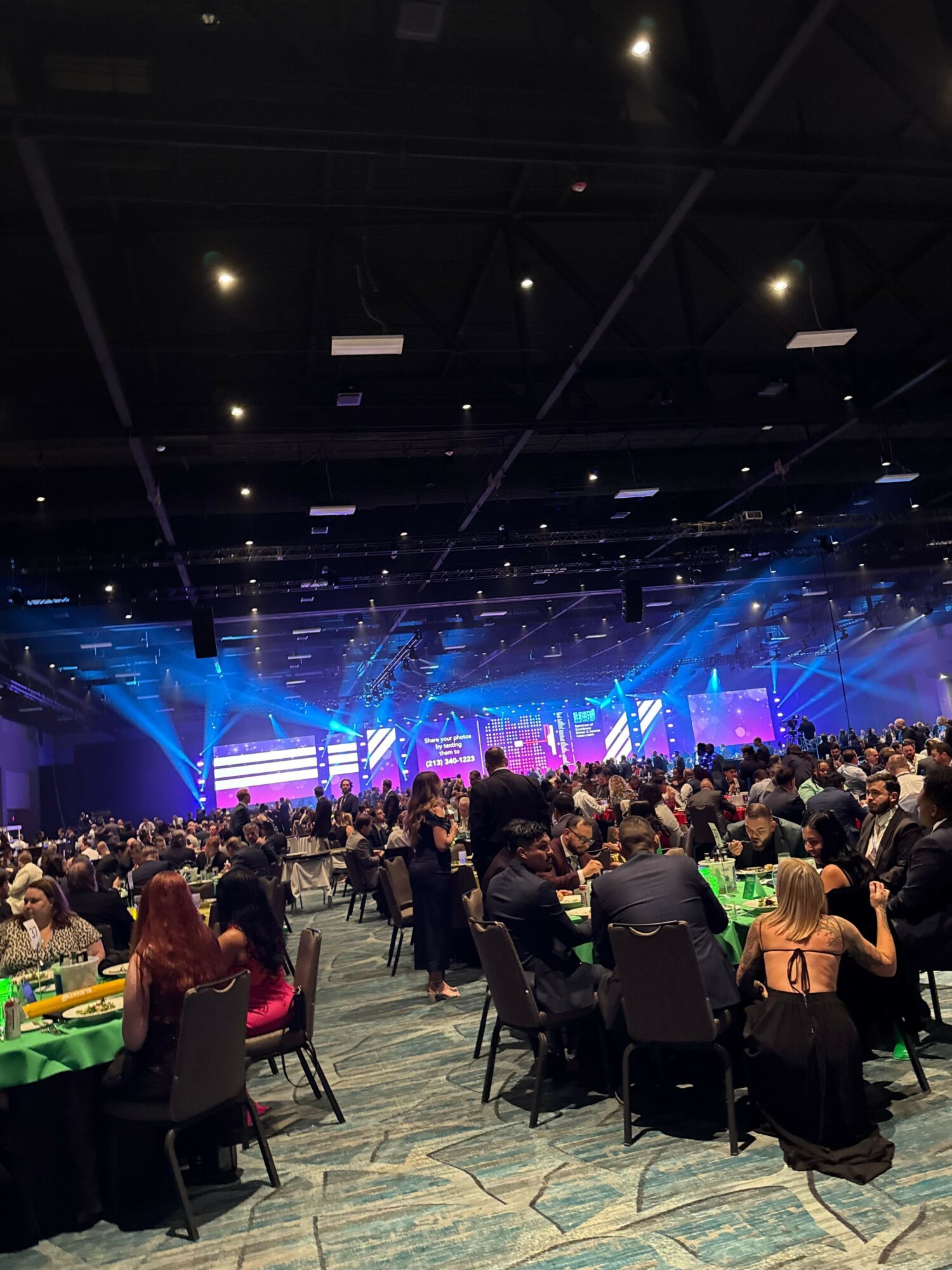 Large indoor event with a stage, audience, and colorful lighting effects, viewed from the side with people seated and standing.