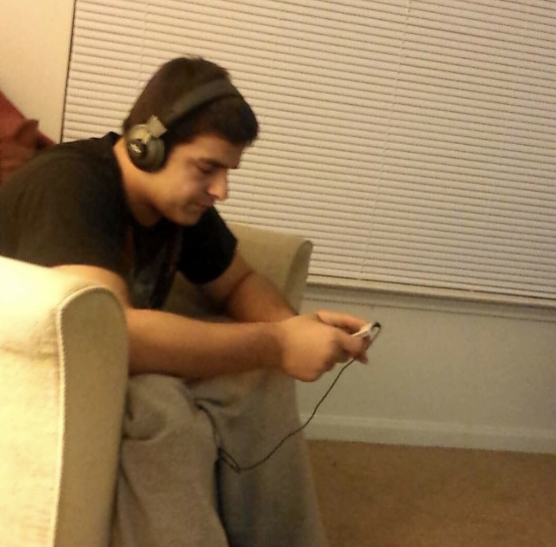 Person sitting on a couch wearing headphones, looking at a phone, with blinds in the background.