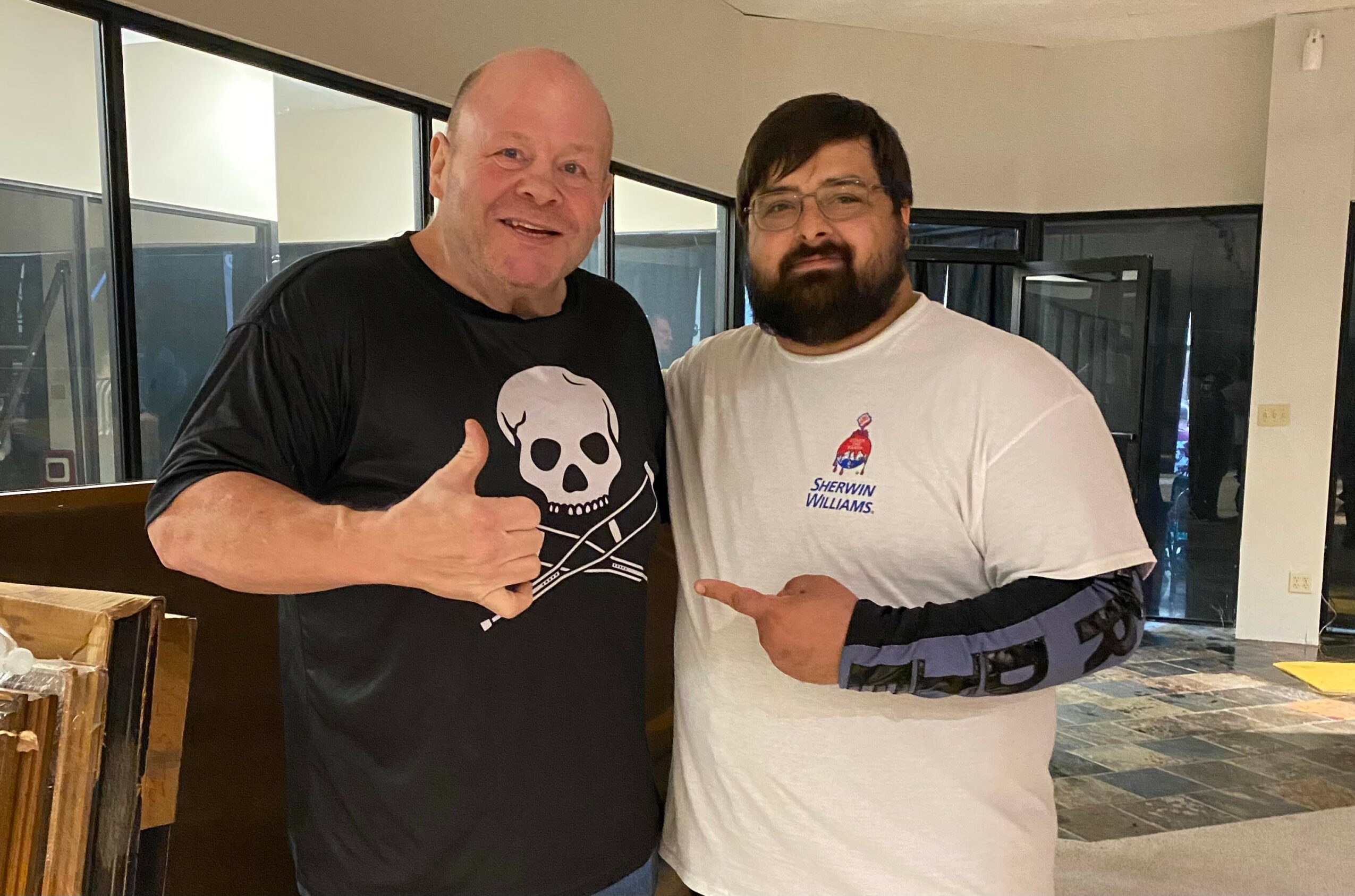 Two men standing indoors, smiling, with large windows behind them, one wearing a black shirt with a skull design, the other in a white shirt with a logo, pointing at each other.