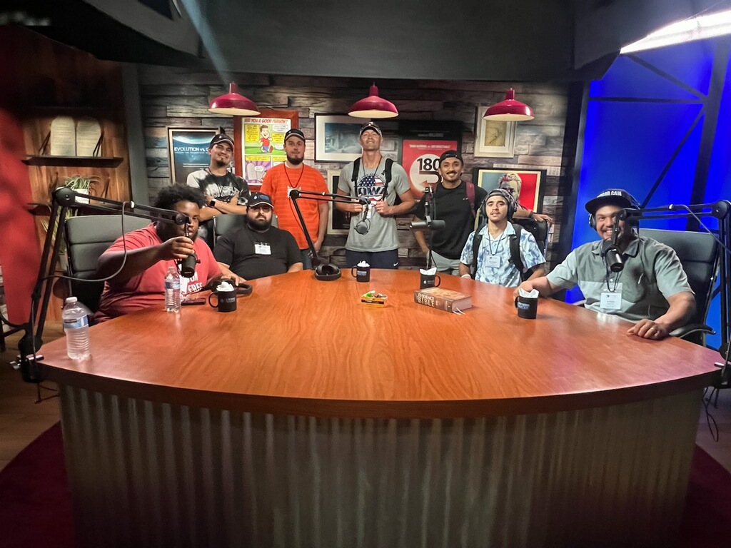 Group of nine people around a large oval table in a studio setting, some holding drinks, with a brick wall background and studio lights.