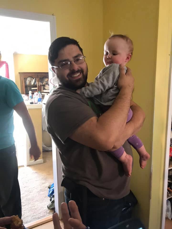 Man holding a baby in a yellow room, smiling, with a window and doorway in the background.