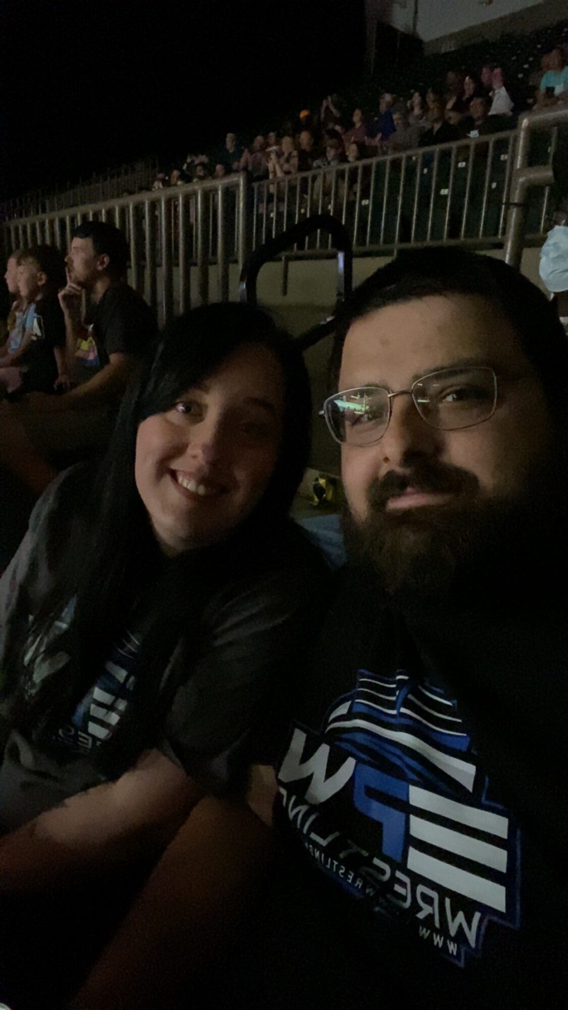 Two people sitting close together at an outdoor event during nighttime, with a crowd in the background.