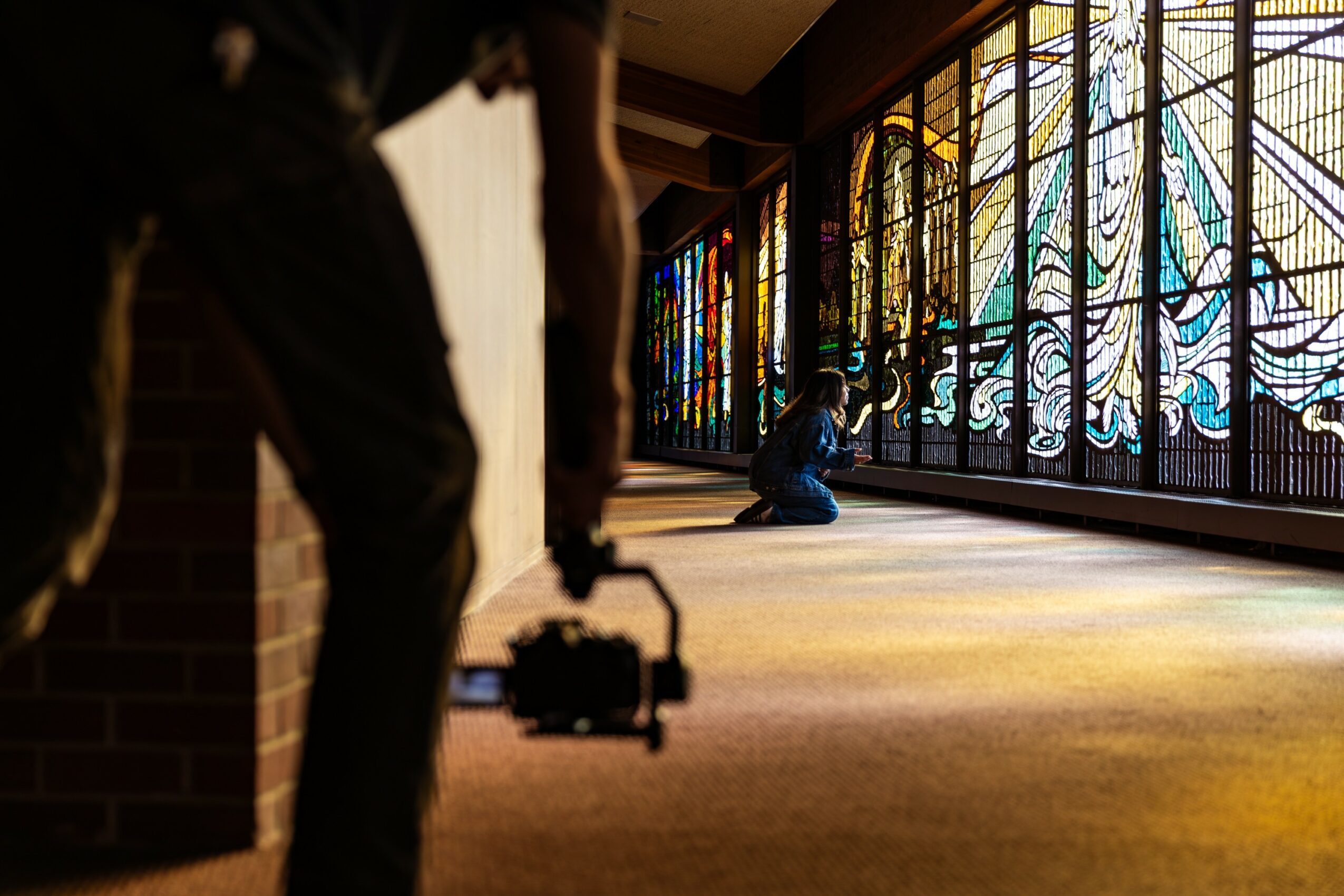 Person holding a camera walking past stained glass windows with a person sitting on the floor nearby, sunlight streaming in.