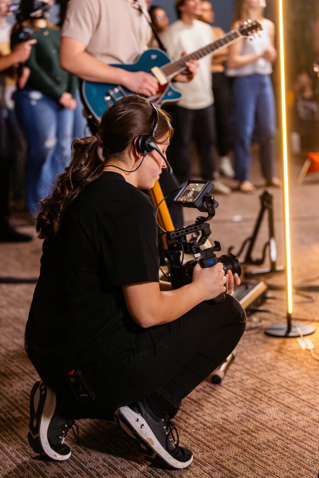 A woman crouches with a camera on a tripod, recording a live music performance indoors. People stand in the background.