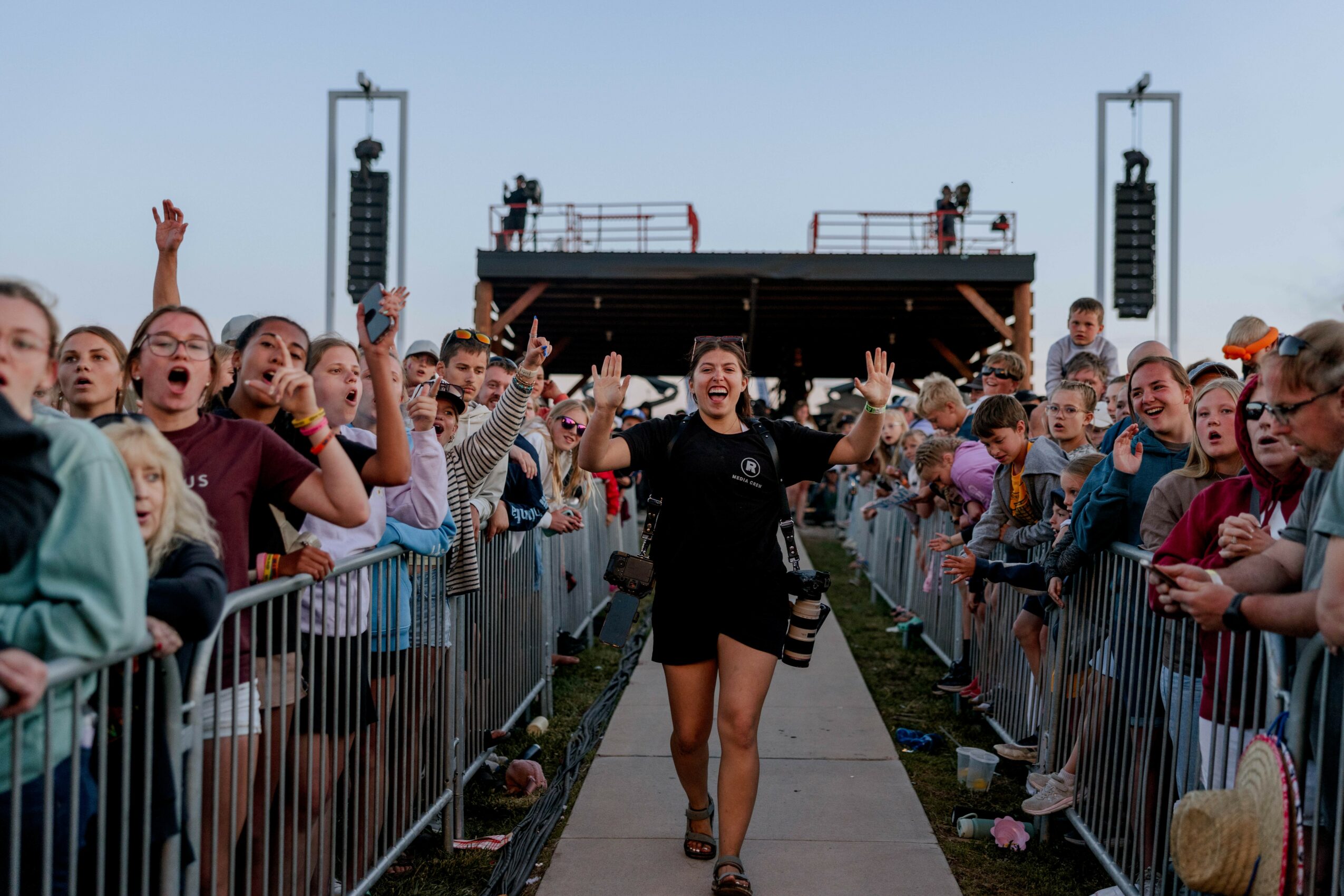 Crowd of people behind barriers at an outdoor event, with a woman walking down a pathway smiling and waving, stage in background.